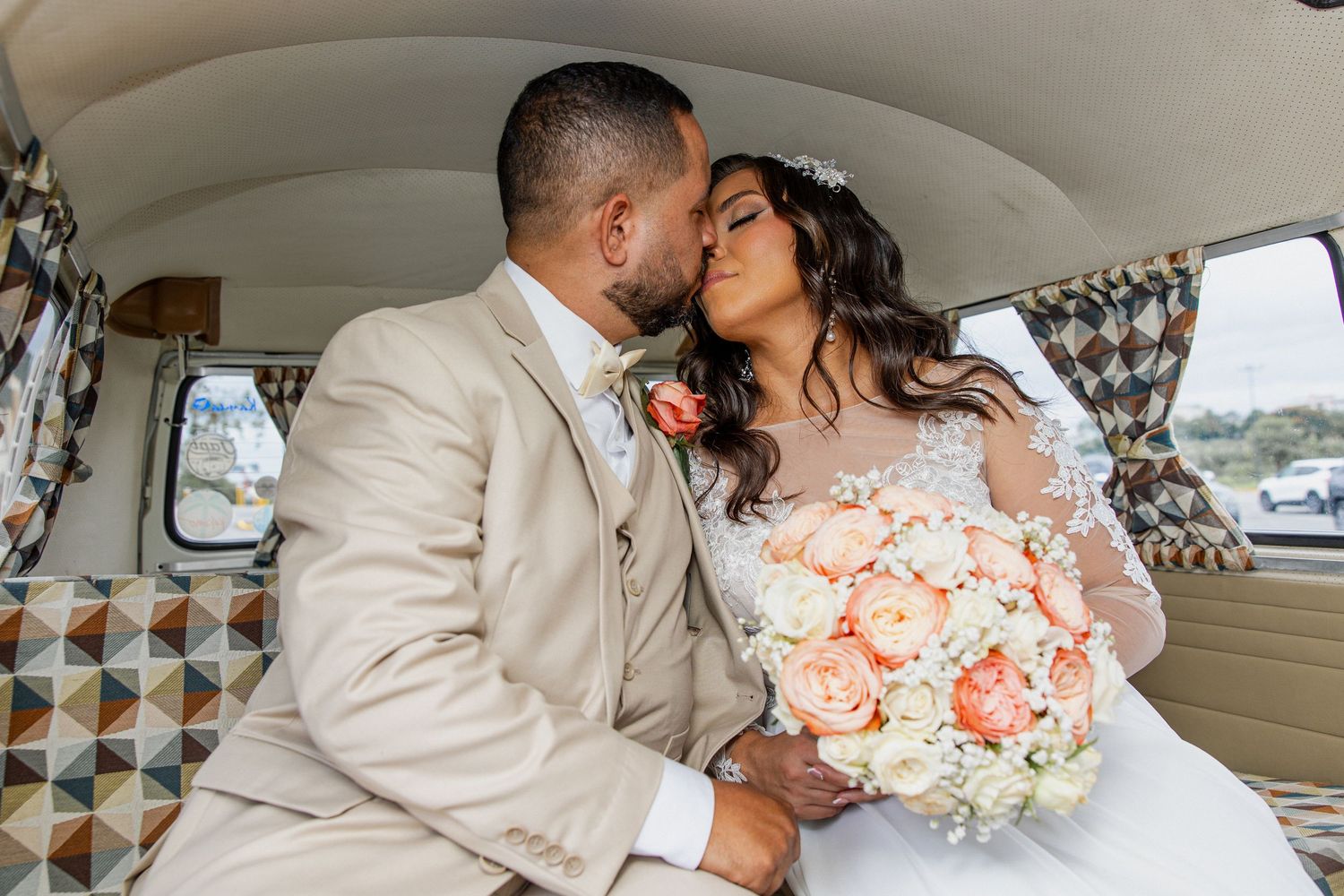 A couple shares romantic moments in a vintage car while dressed in wedding attire with peach and white floral bouquet.