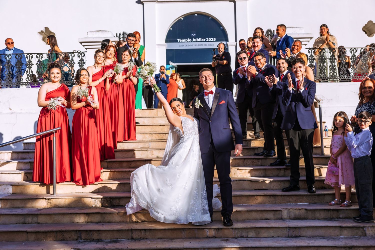 Joyful newlyweds celebrate on church steps surrounded by bridesmaids in red dresses and cheering guests at a wedding ceremony.