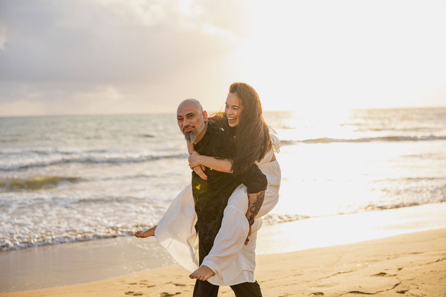 A couple shares a joyful piggyback moment on the beach during golden hour as waves crash behind them.
