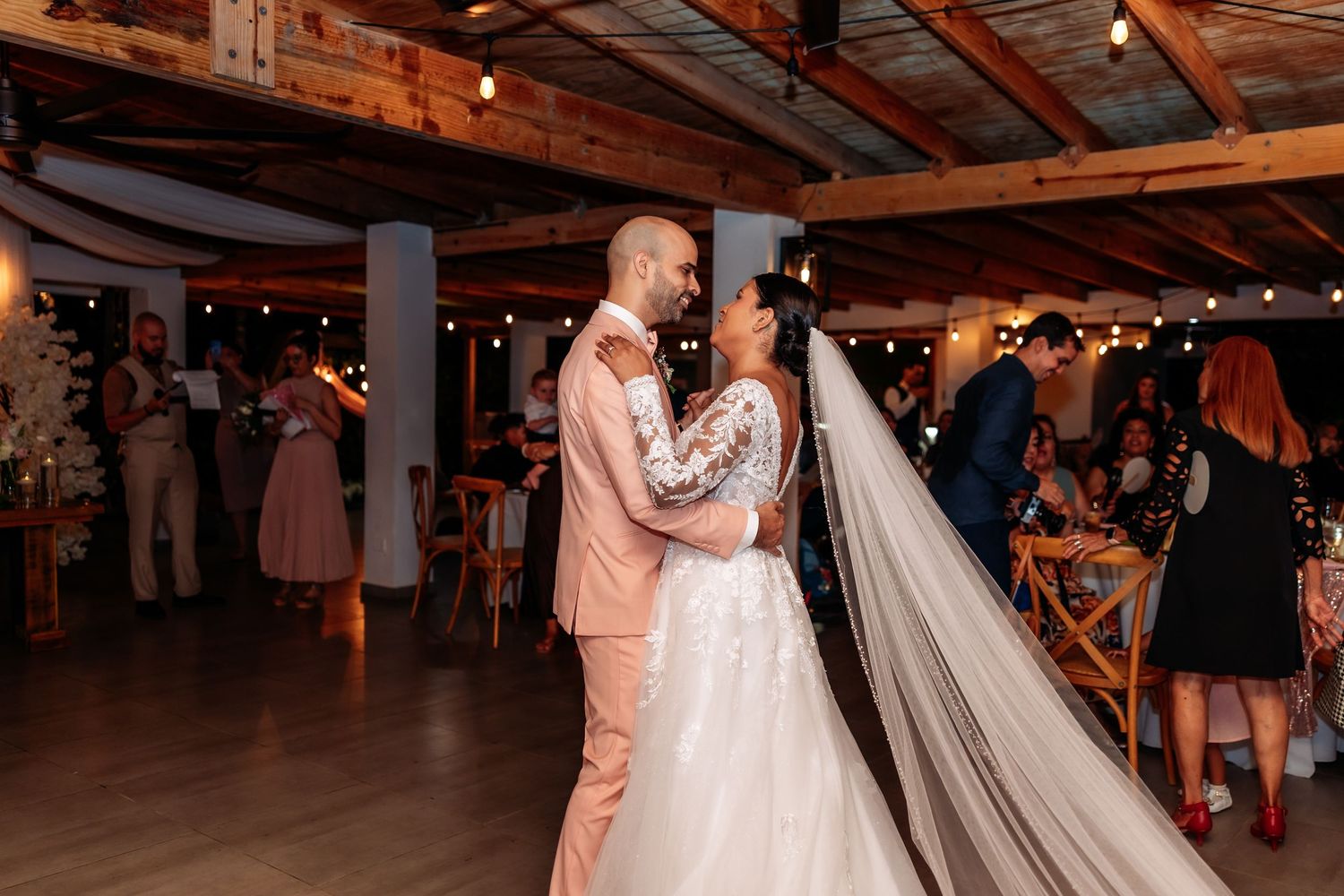 A couple shares their first dance at a rustic barn wedding reception with string lights overhead.