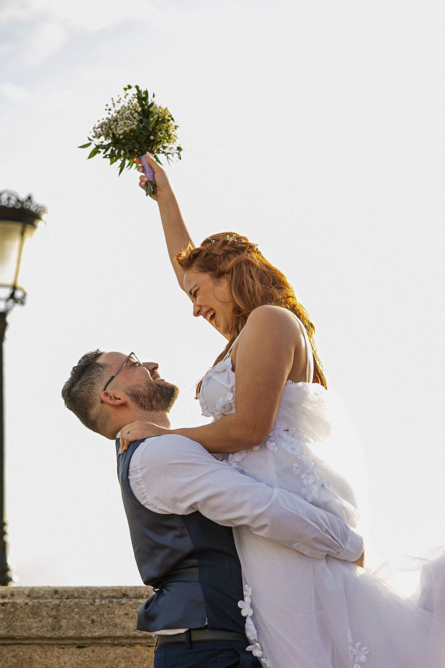 Groom lifting smiling bride in white dress holding bouquet aloft outdoors on their wedding day.