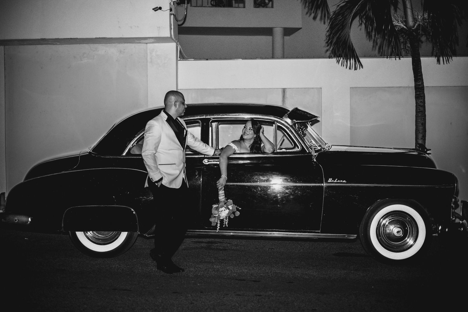 Couple posing with a vintage black car at night in a classic black and white wedding photo.