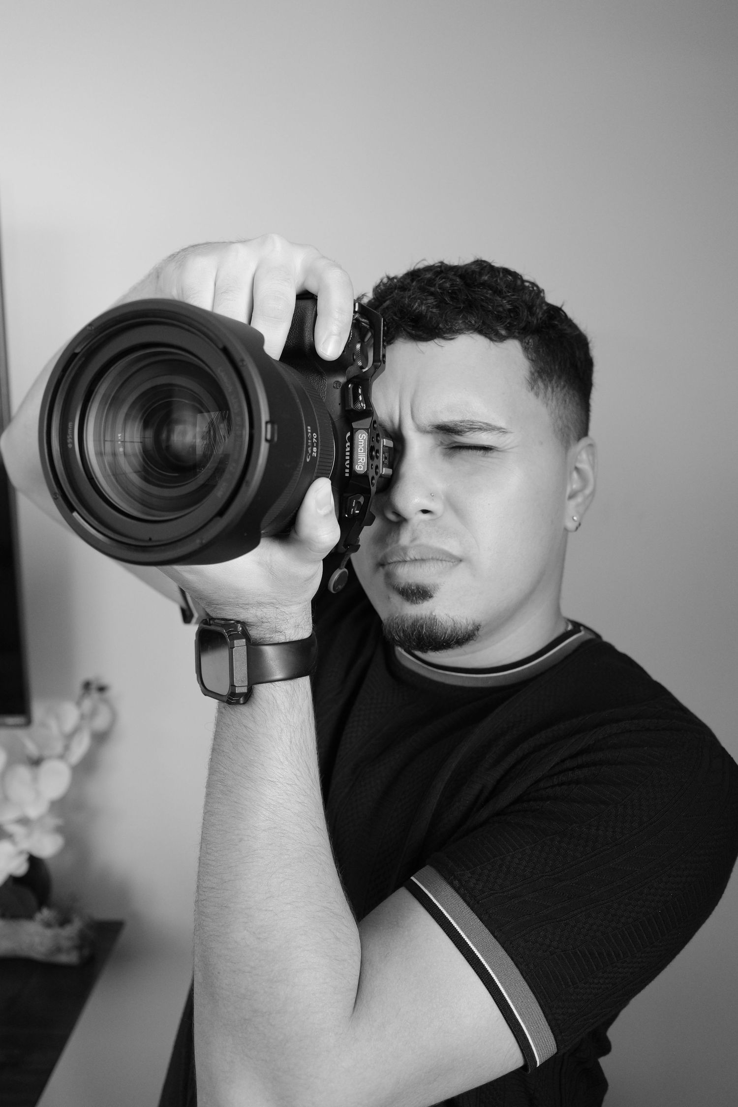 Black and white photo of a young man holding a DSLR camera up to his eye, wearing a black t-shirt.