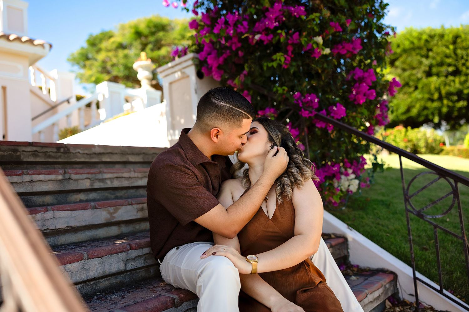 A romantic couple embraces on stone steps surrounded by vibrant pink bougainvillea flowers in a garden setting.