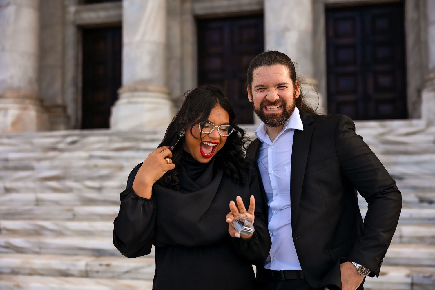 Two people laughing and posing together on stone steps outside a building with columns.