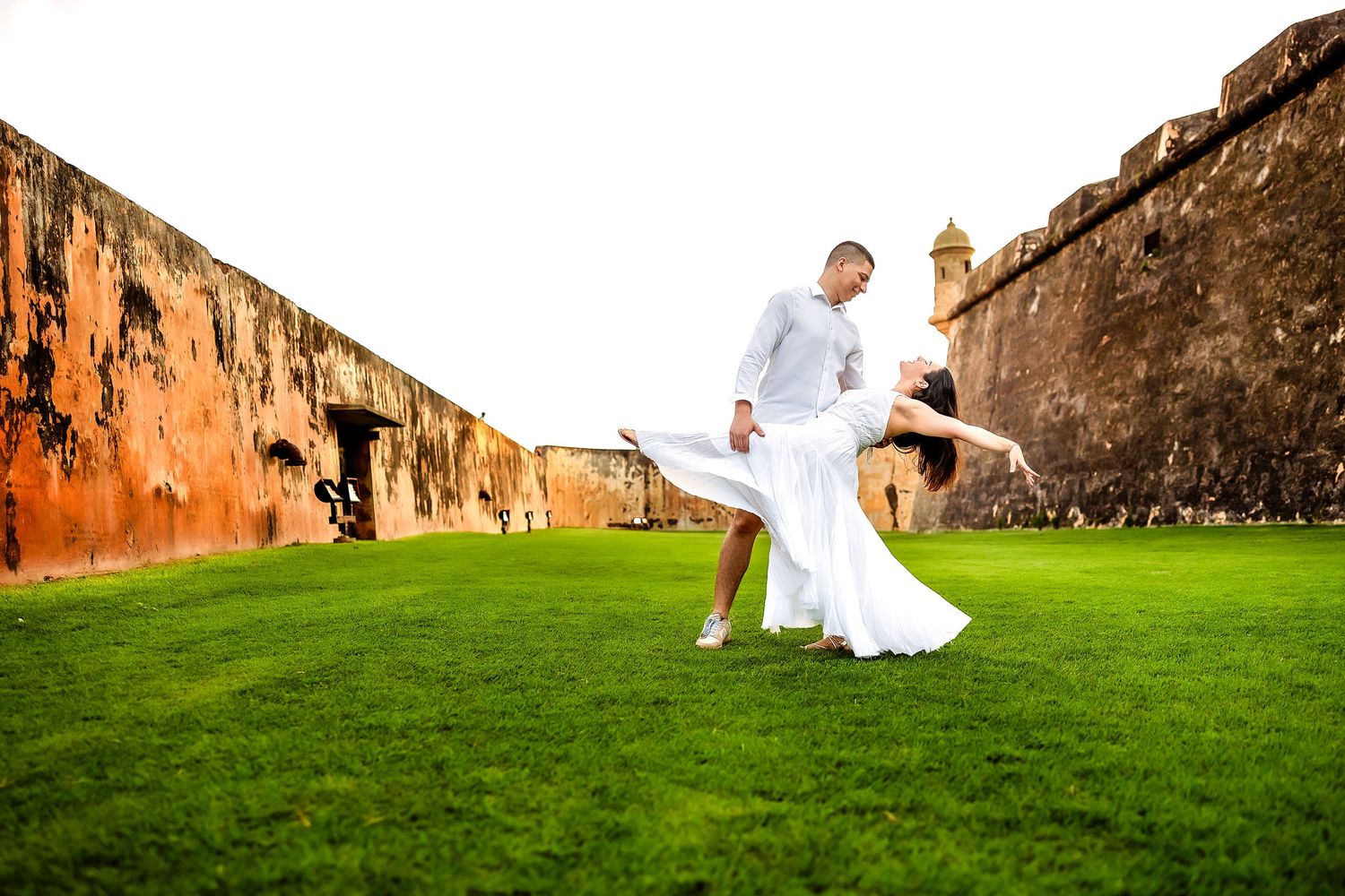 A couple in wedding attire walks along a historic stone wall and grassy path in a romantic outdoor setting.