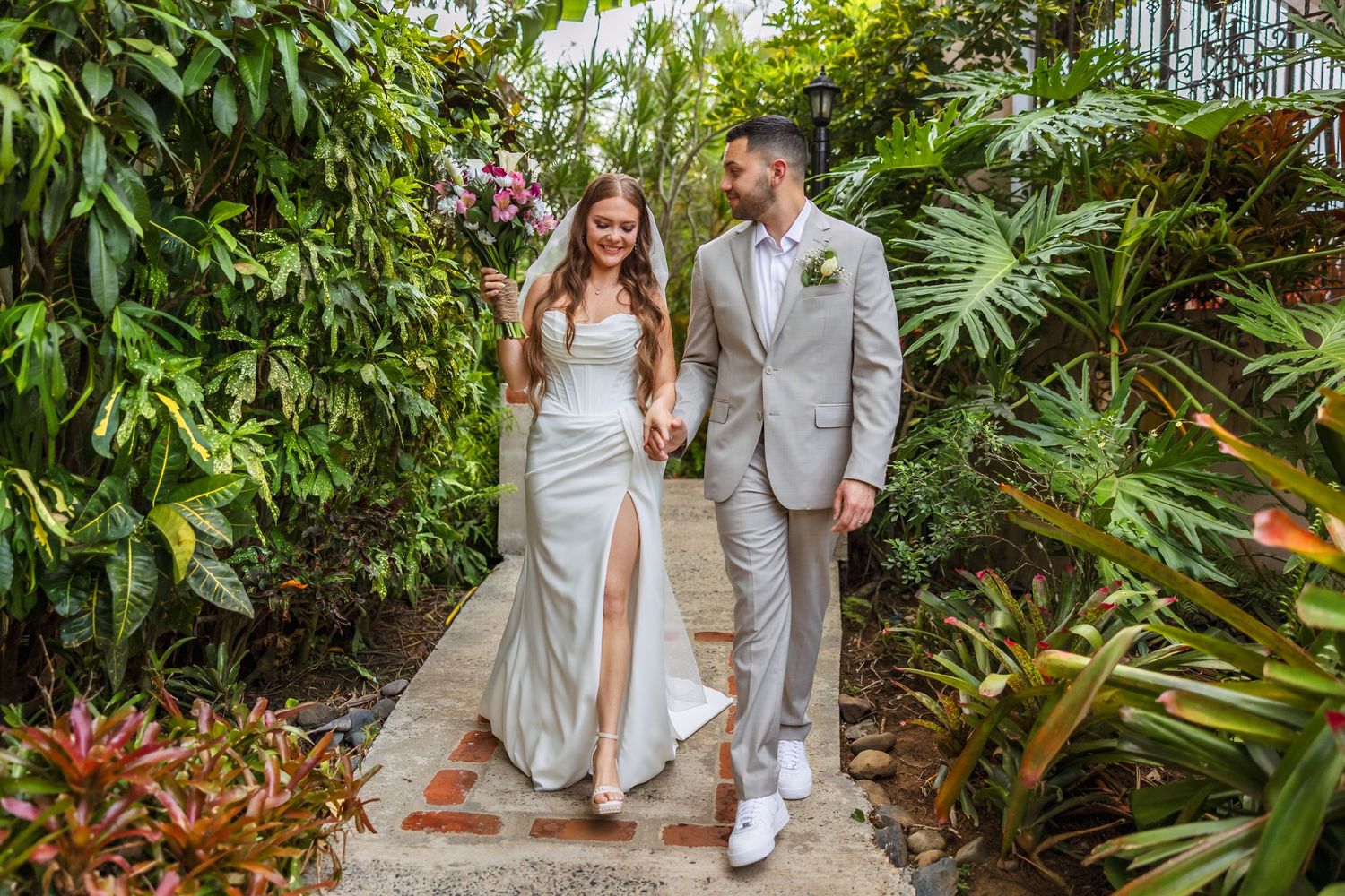 Bride and groom walk hand-in-hand through a lush tropical garden path on their wedding day.