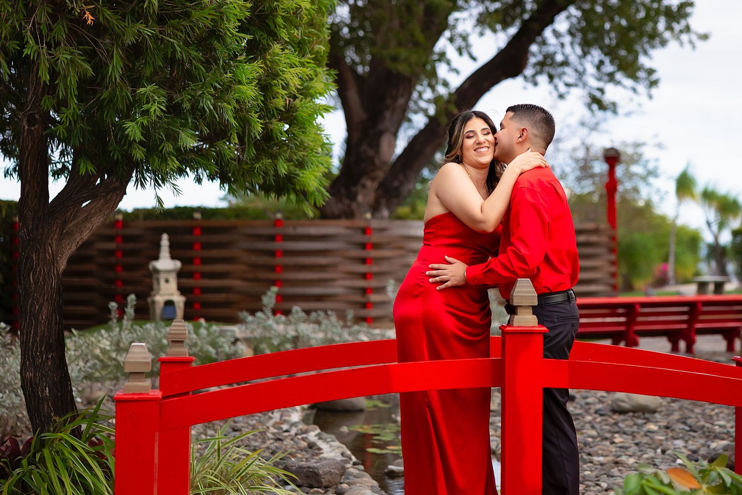 Couple in matching red outfits embracing on a red bridge in a Japanese garden setting.