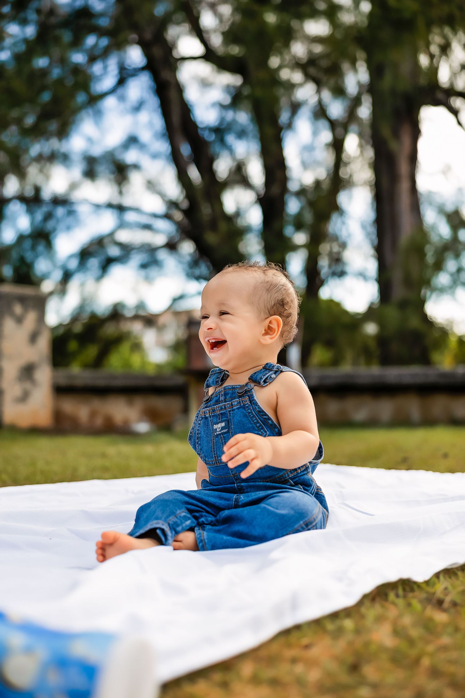 Happy baby in denim overalls sitting on white blanket outdoors, laughing joyfully in a sunny park setting.