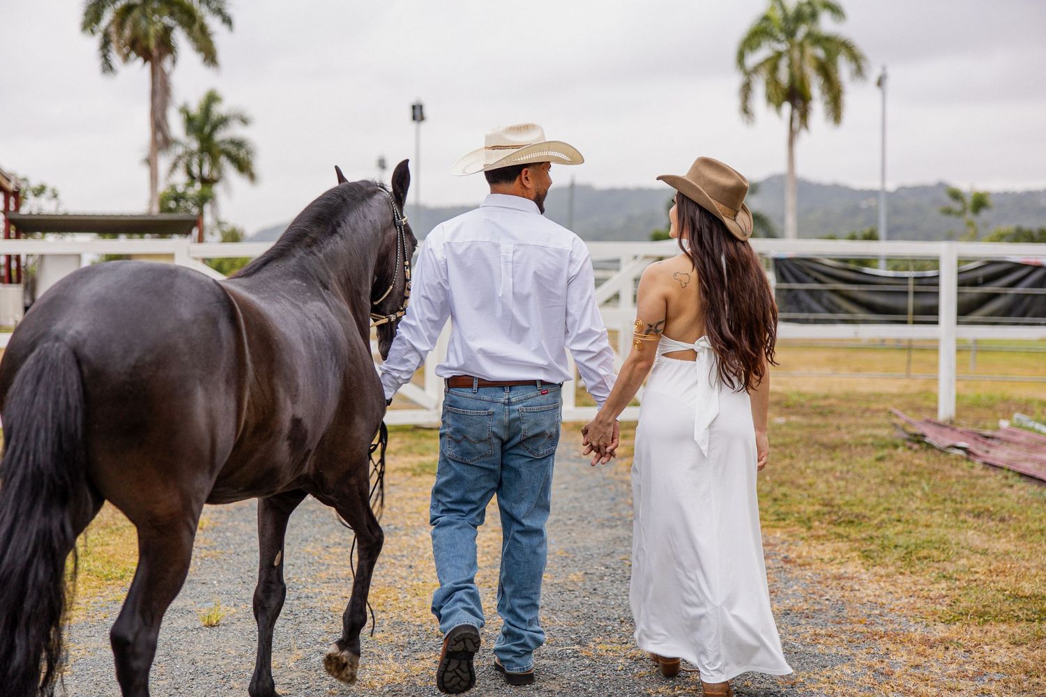 Couple in western attire walking hand in hand with a black horse at a ranch with palm trees.