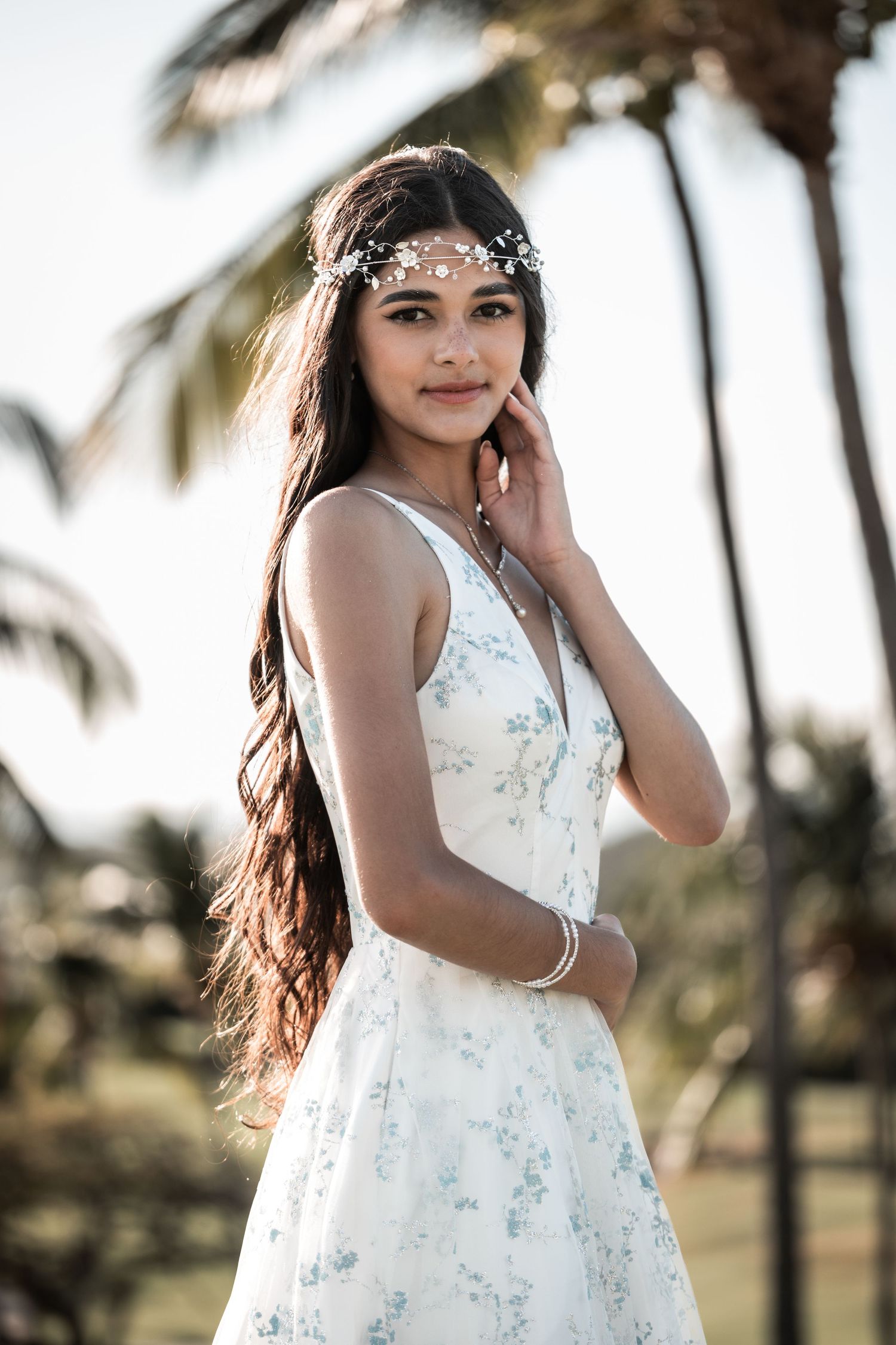 Young woman in white floral dress and pearl headpiece poses gracefully outdoors near palm trees.