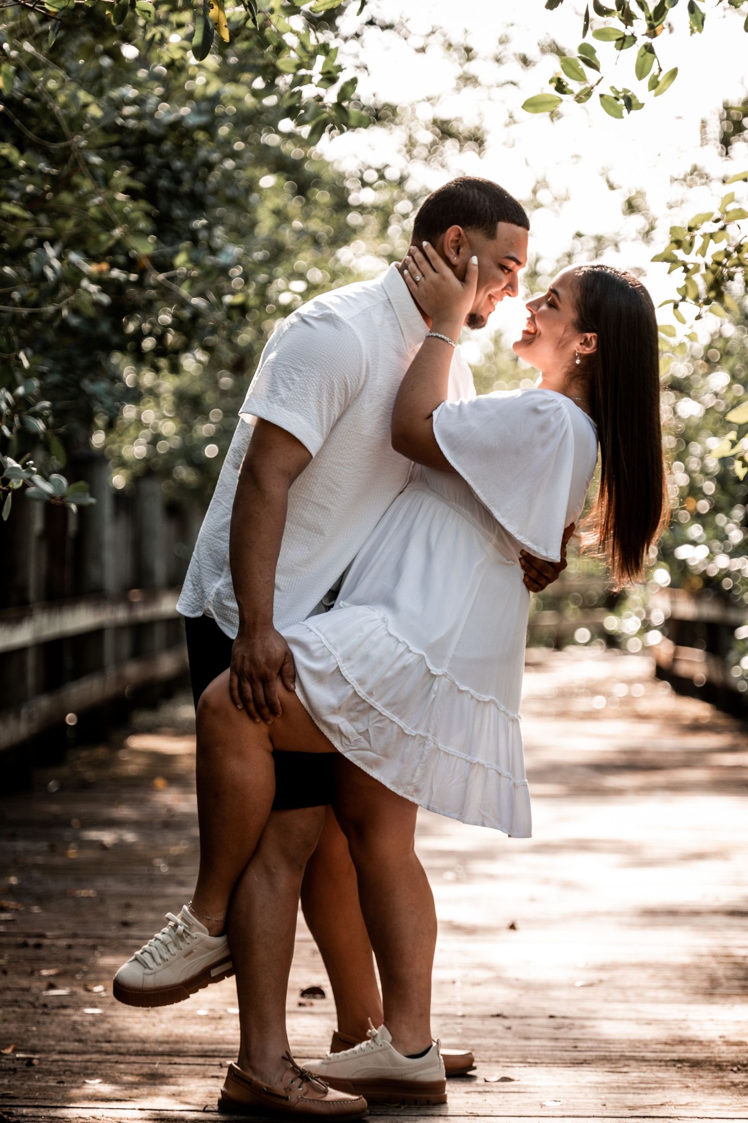 Couple in matching white outfits sharing a romantic moment on a sunlit wooden path surrounded by lush green trees.