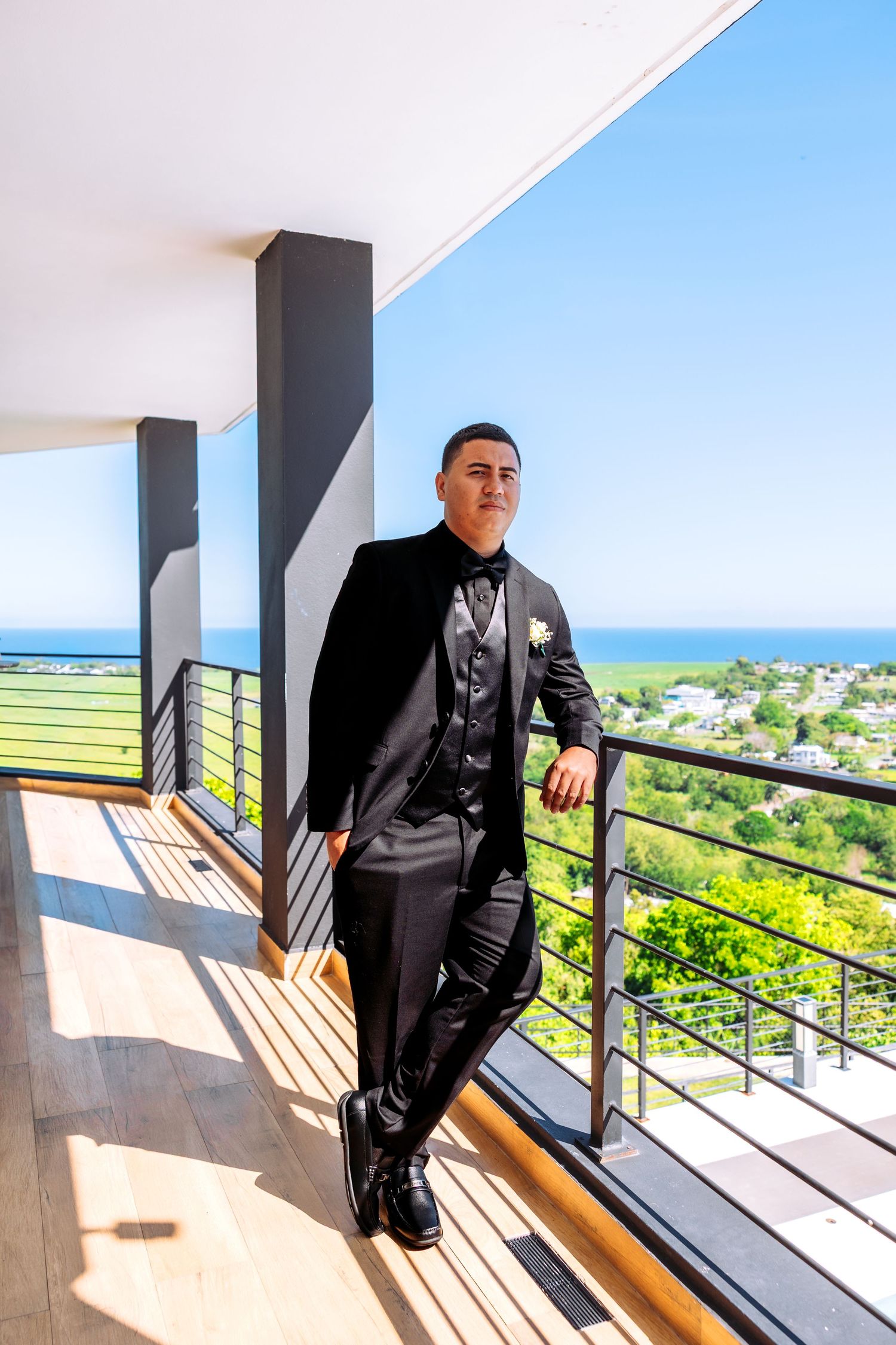 Groom in black tuxedo with boutonniere poses on sunny balcony overlooking ocean and green landscape.