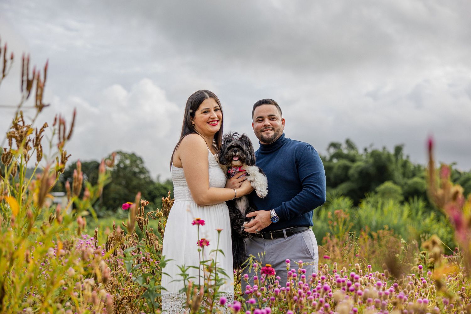 Couple holding a black dog in a colorful flower field with cloudy skies in the background.