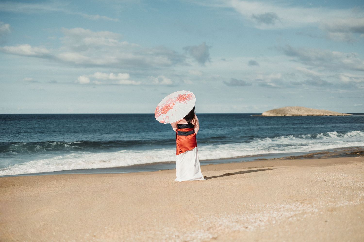 A figure with a pink parasol stands on a beach against an ocean backdrop.