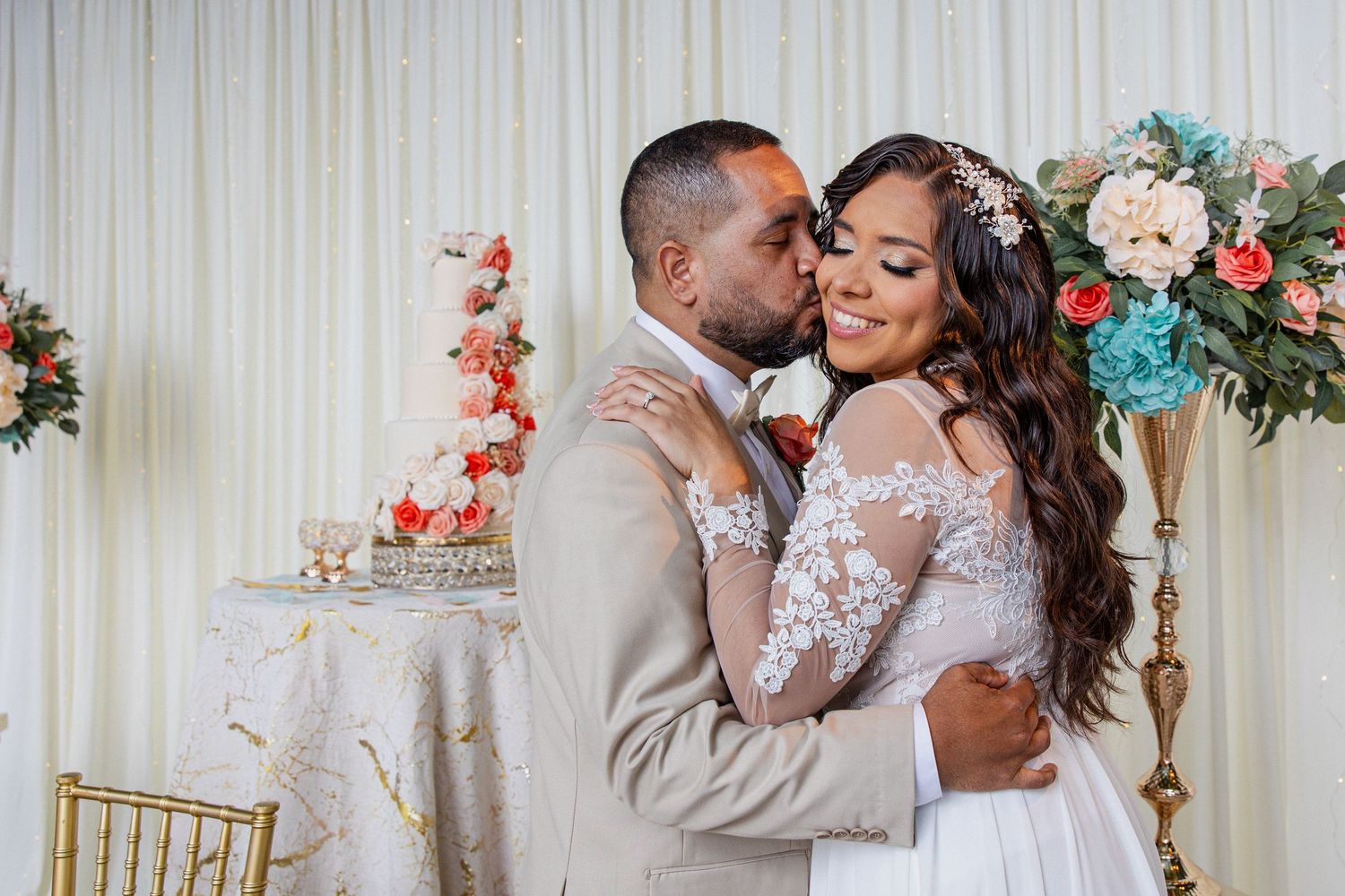 A groom in a tan suit kisses his bride in a lace wedding gown near a floral cake at their elegant reception.