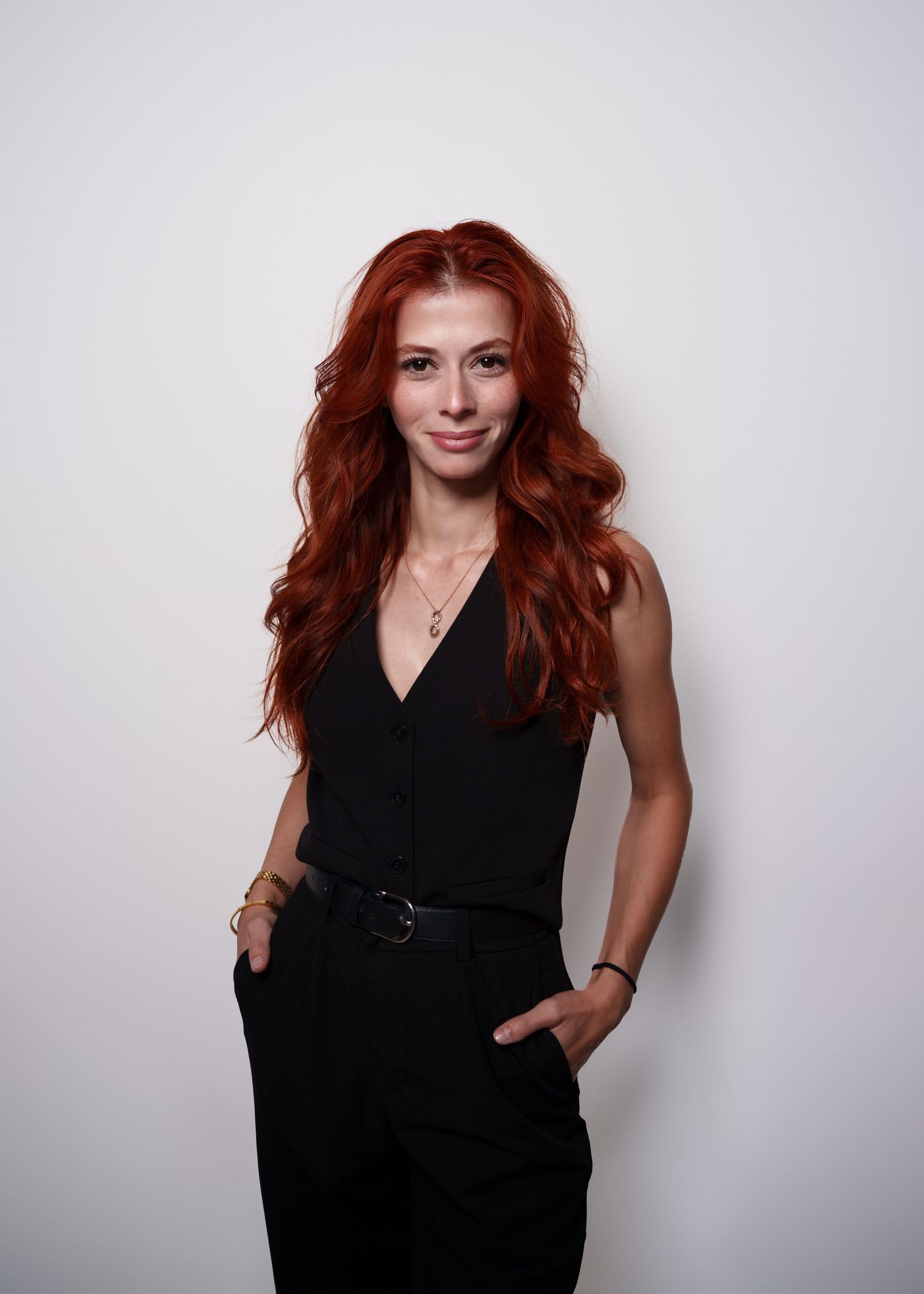 A woman with vibrant red hair wearing a sleek black dress poses confidently against a white background.