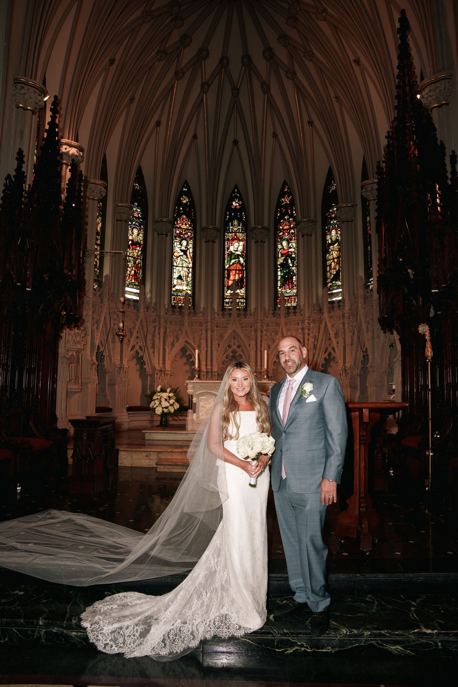 A couple poses together in a beautiful gothic church with stained glass windows and a flowing lace wedding dress train. wedding new york
