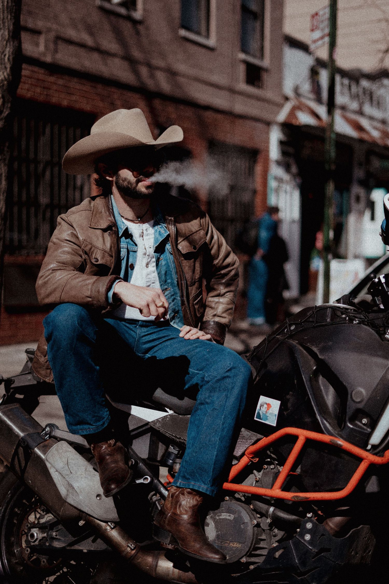 Person in cowboy hat and brown jacket sitting on ATV while smoking on city street. lower manhattan soho