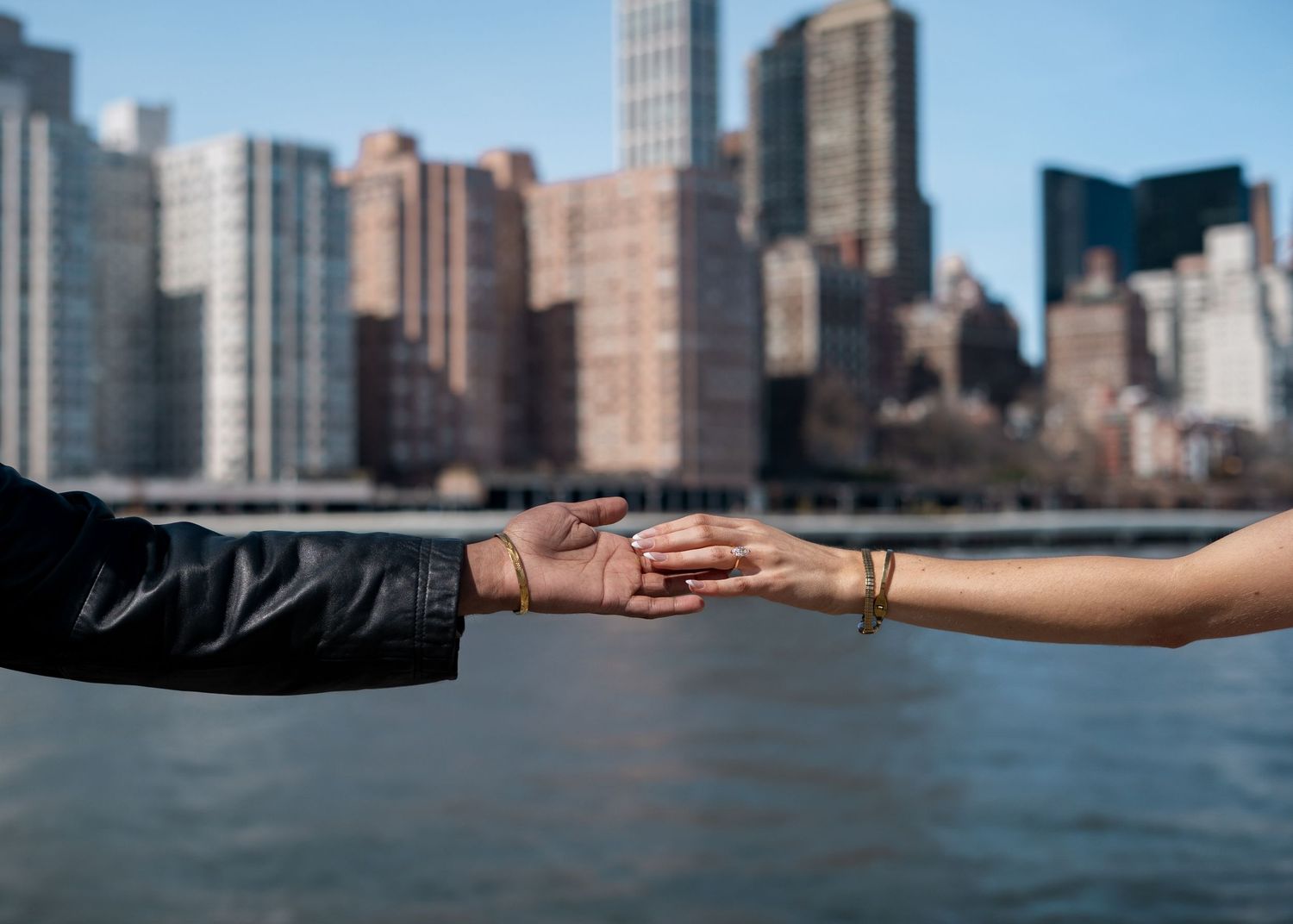 Two hands reaching towards each other with New York City skyline in the background across the water.