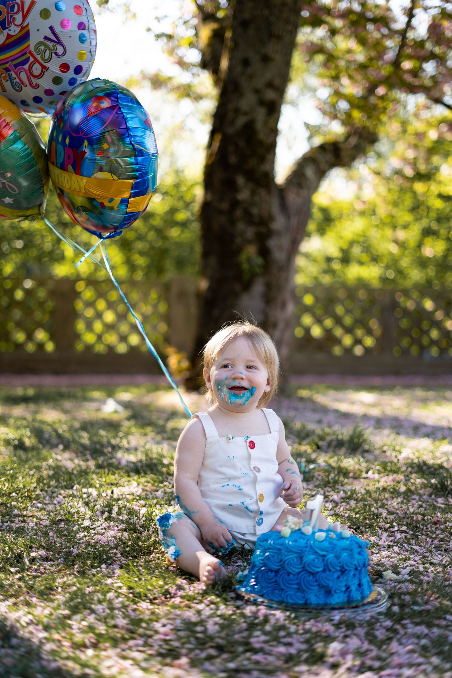 A small child sits in a blue tutu outdoors with a birthday balloon and scattered leaves on the ground. cake smash