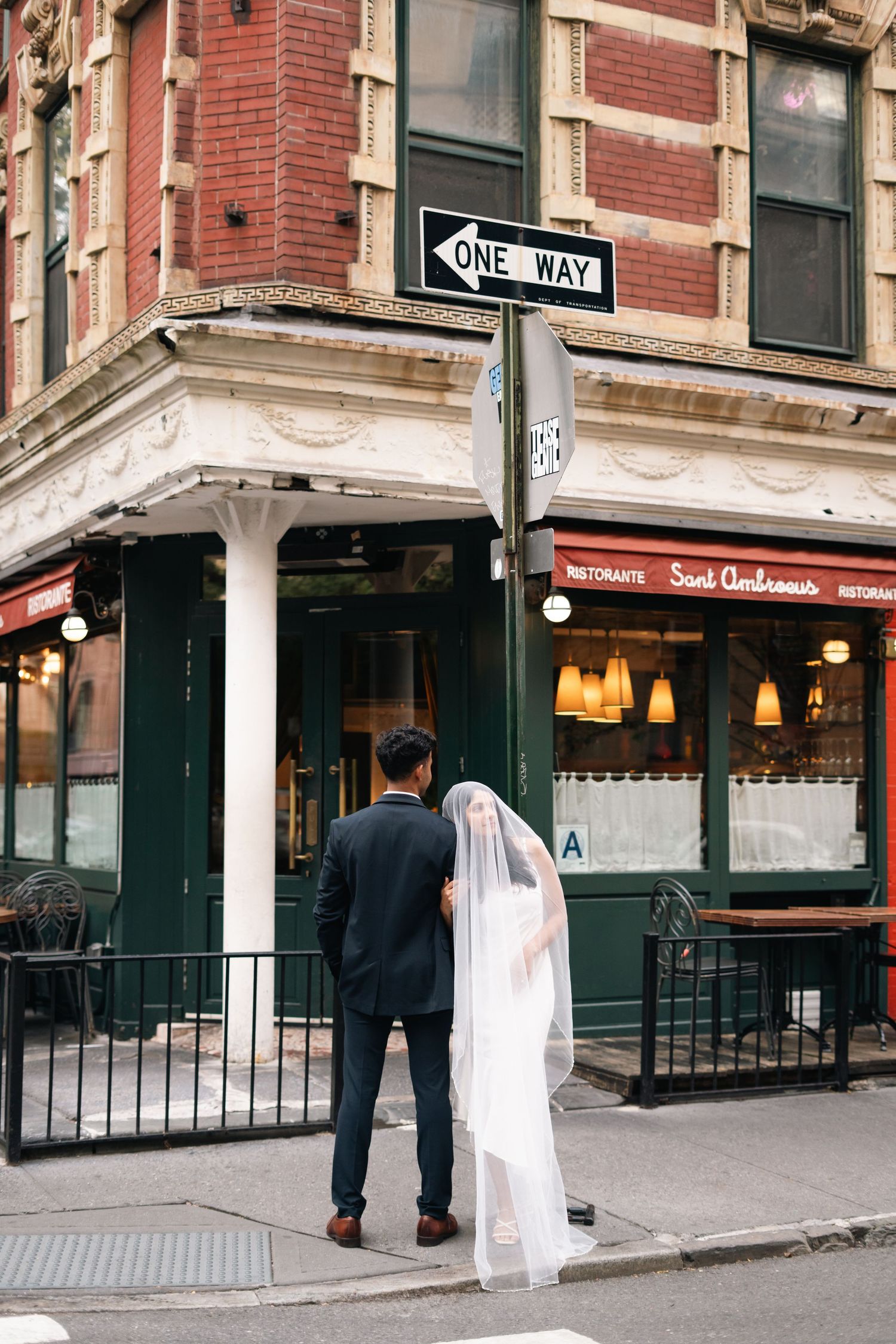 A couple stands on a street corner beneath a one-way sign outside a brick building with a restaurant storefront. soho wedding engagement ny