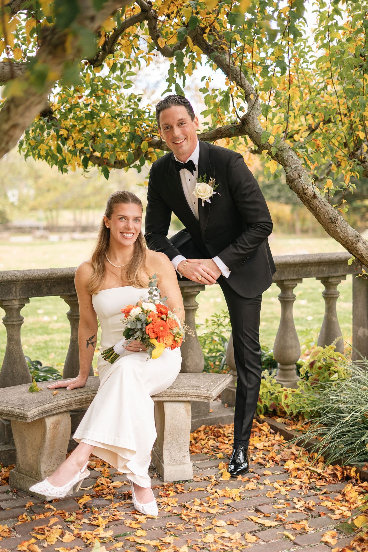 Newlywed couple poses playfully in autumn setting with fallen leaves and stone balustrade under yellow trees. in new york, connecticut, nj