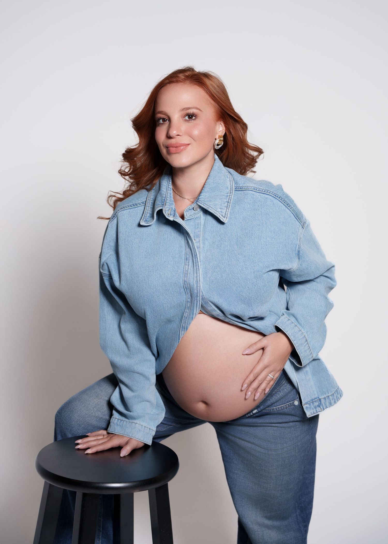 Pregnant woman in light denim jacket sits on black stool showing her baby bump against white background in new york