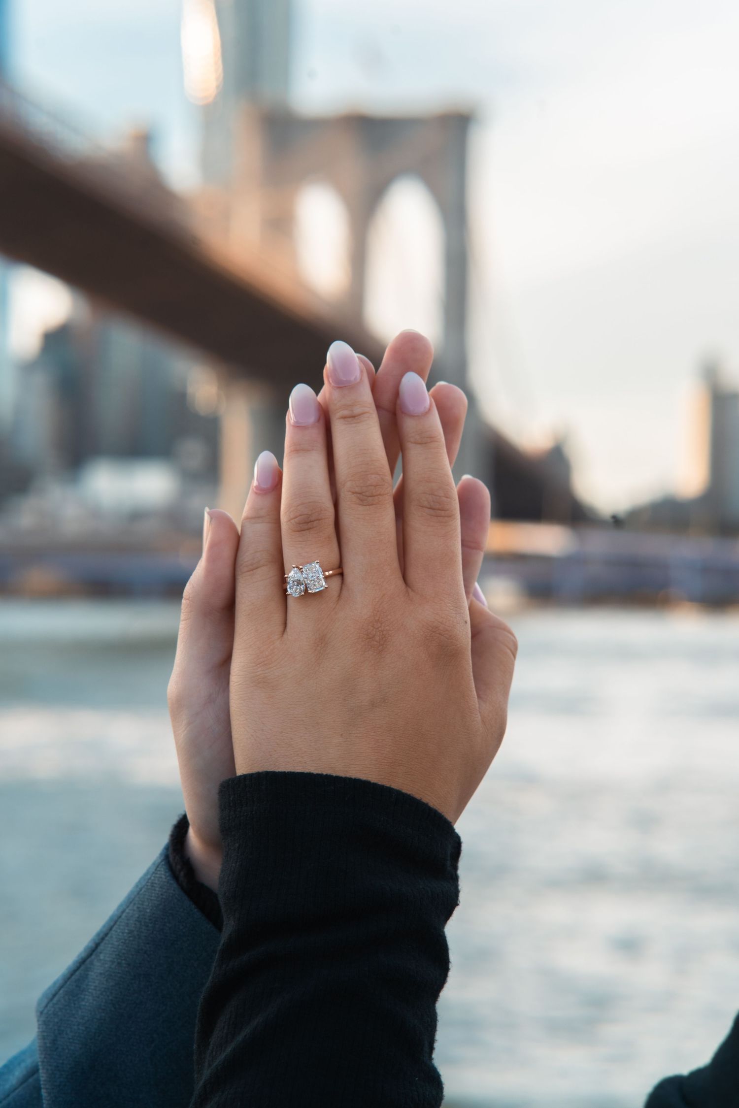 A diamond engagement ring sparkles on a hand against a blurred waterfront cityscape background. brooklyn bridge proposal