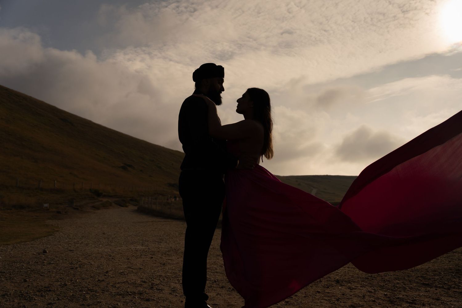 Silhouetted couple embracing on hilltop at sunset with flowing red fabric creating dramatic movement in the wind.
