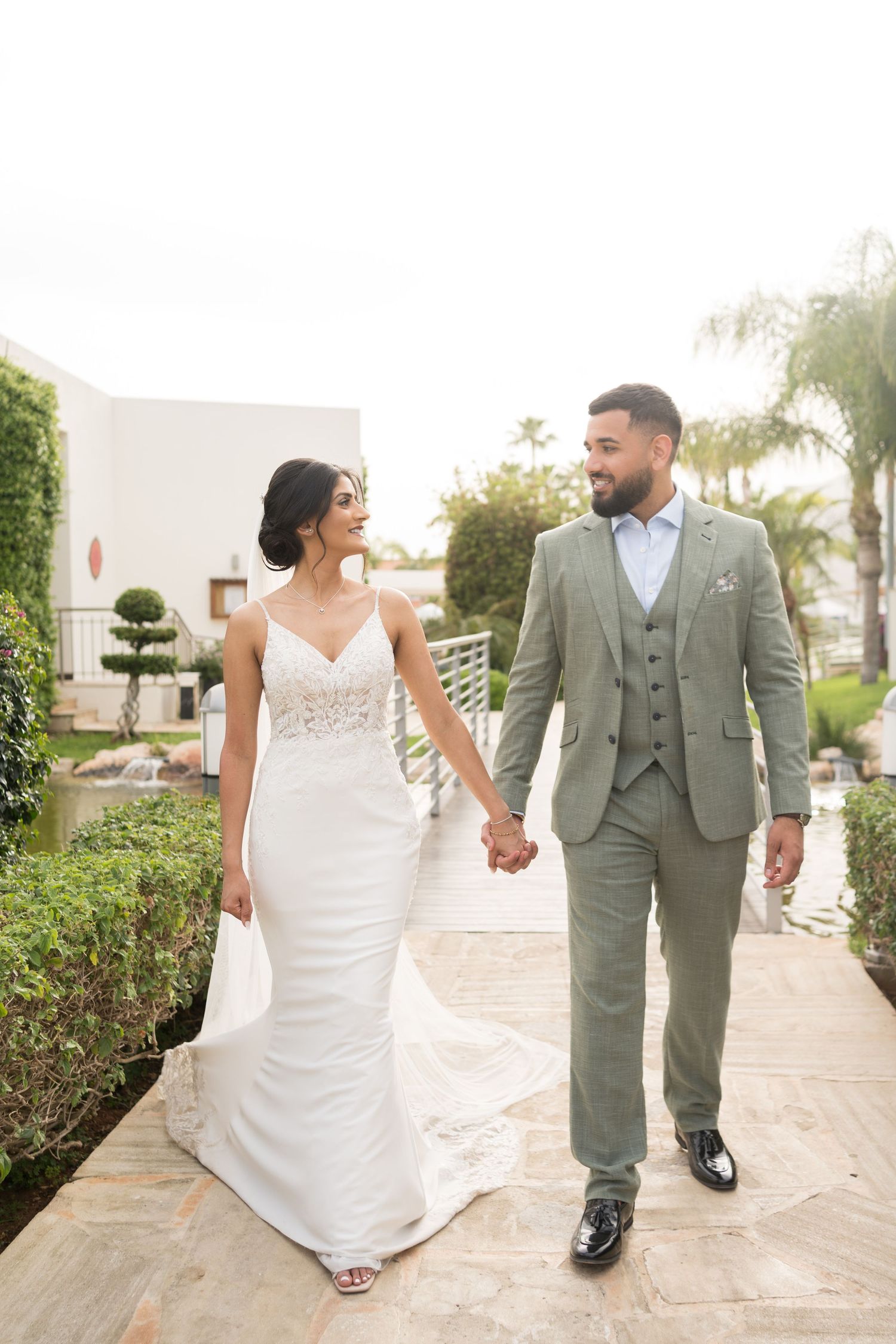 Wedding couple walking hand in hand on garden path, bride in white gown and groom in light grey suit.
