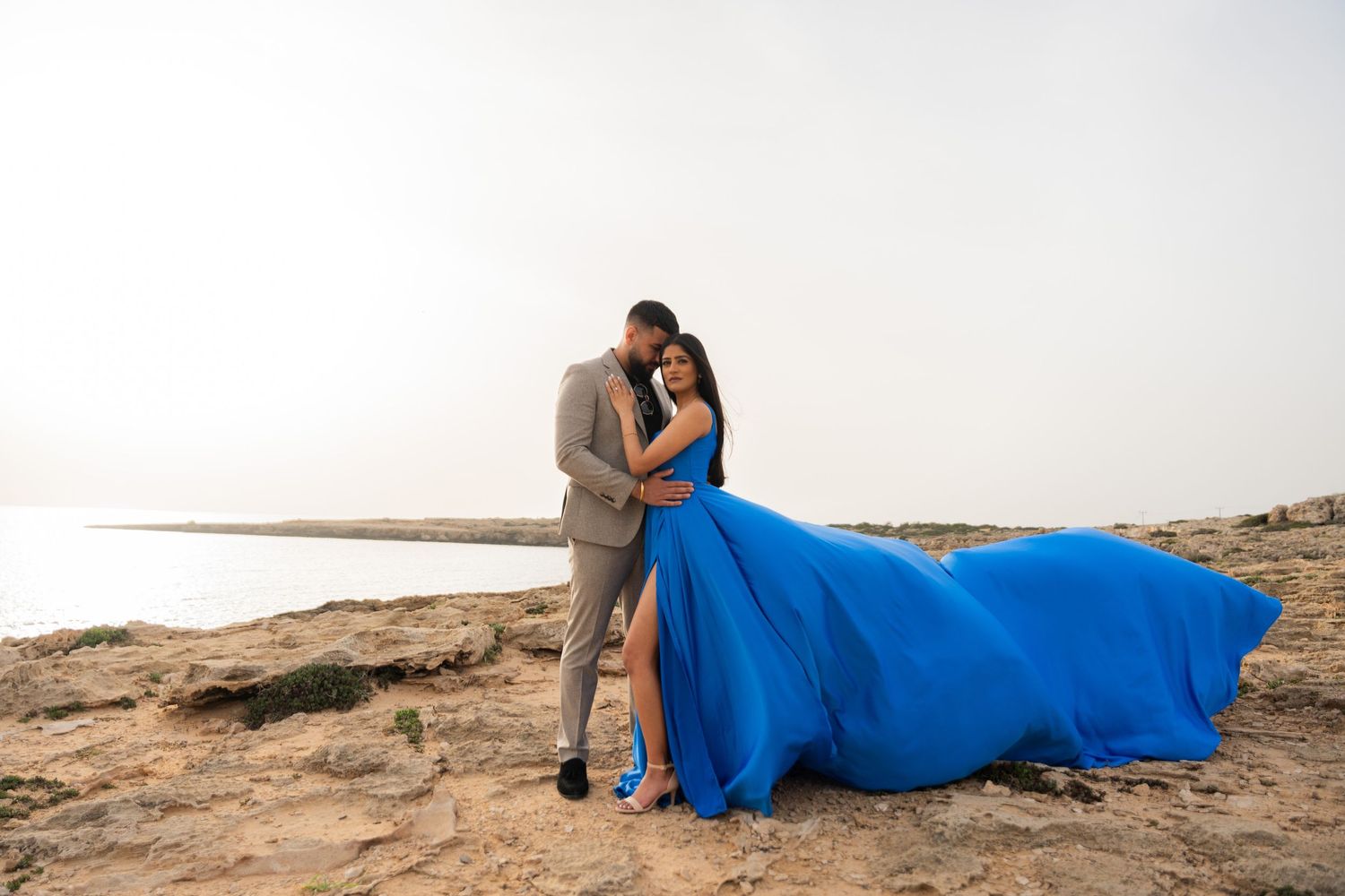Couple embracing on beach with dramatic flowing blue dress in gentle sunlight.