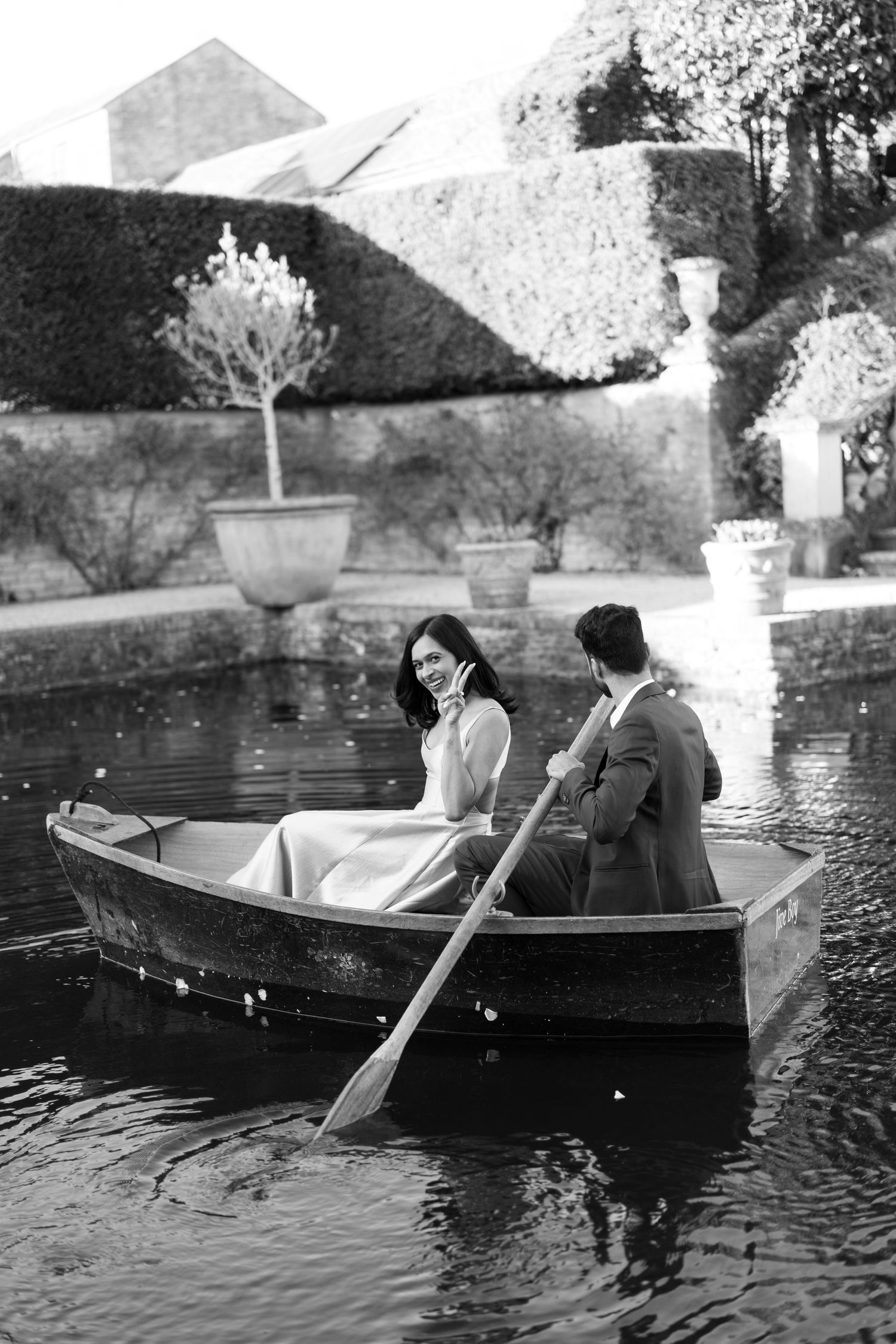 A romantic boat ride scene showing a couple in a small wooden rowboat on a tranquil pond surrounded by garden walls.