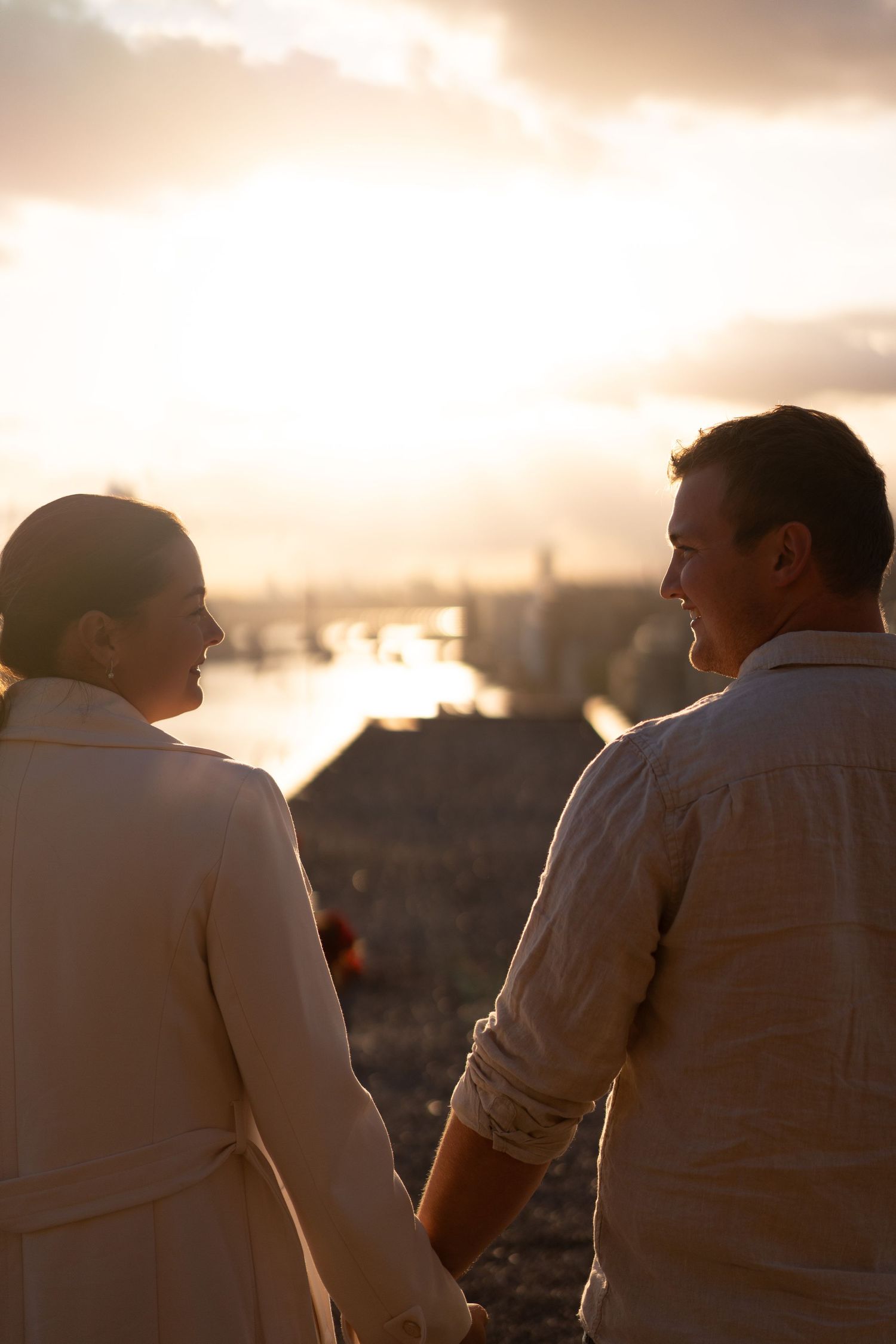 A couple stands together holding hands while watching a romantic sunset over a cityscape.