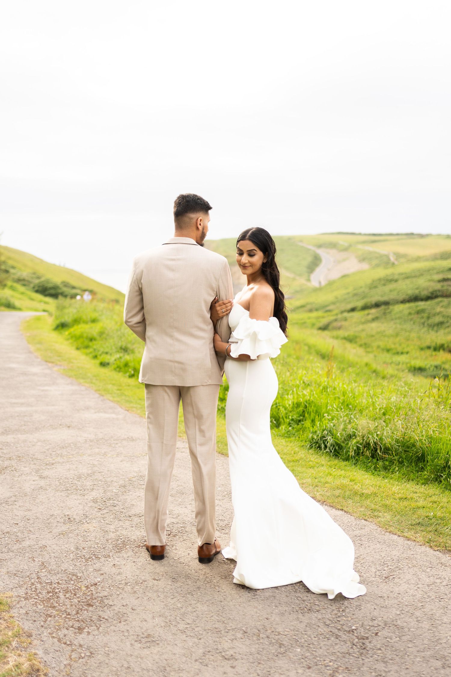 Couple in wedding attire walking along a scenic coastal path with rolling green hills.