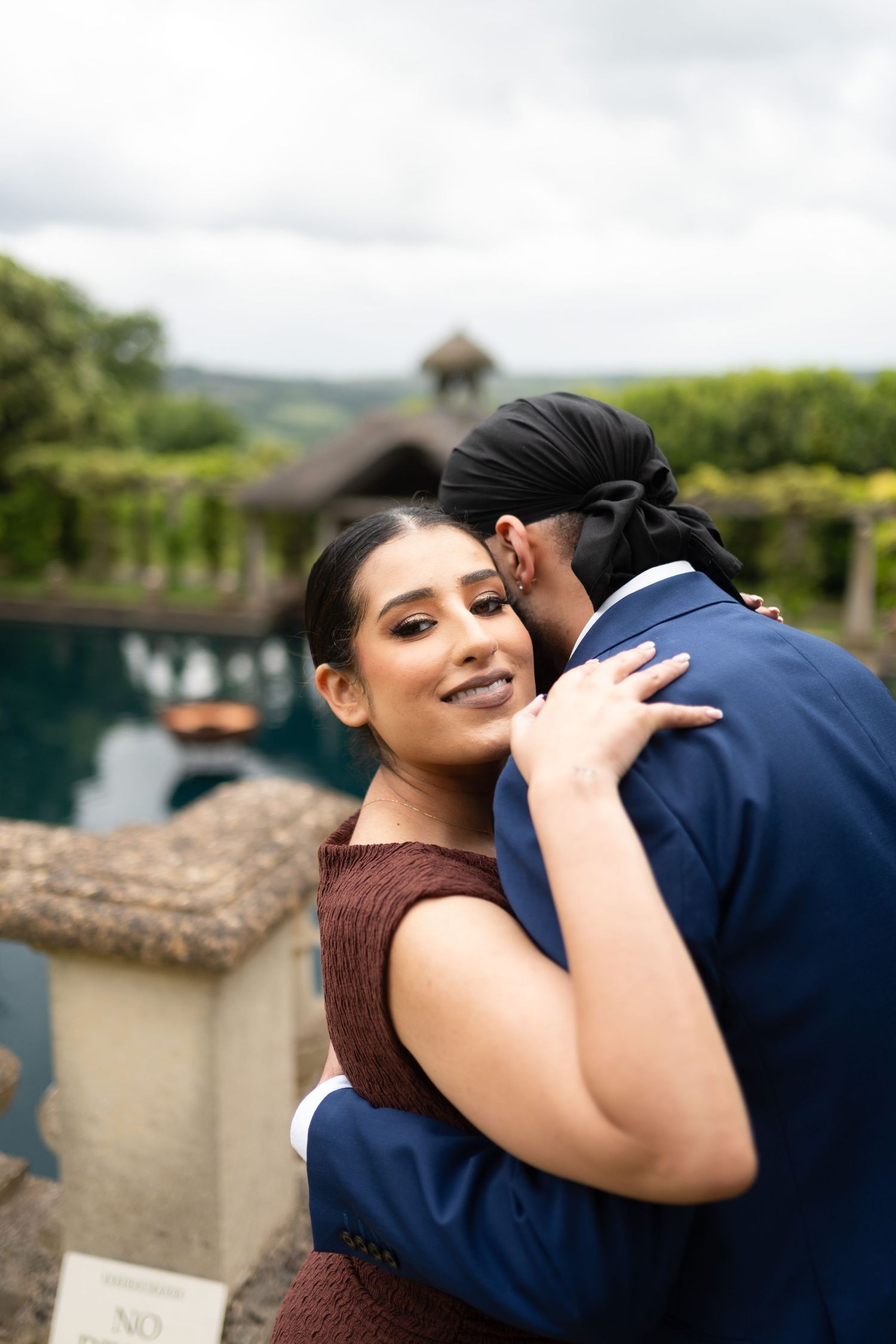 A romantic couple embraces at an elegant venue with a reflection pool and gazebo in the background.