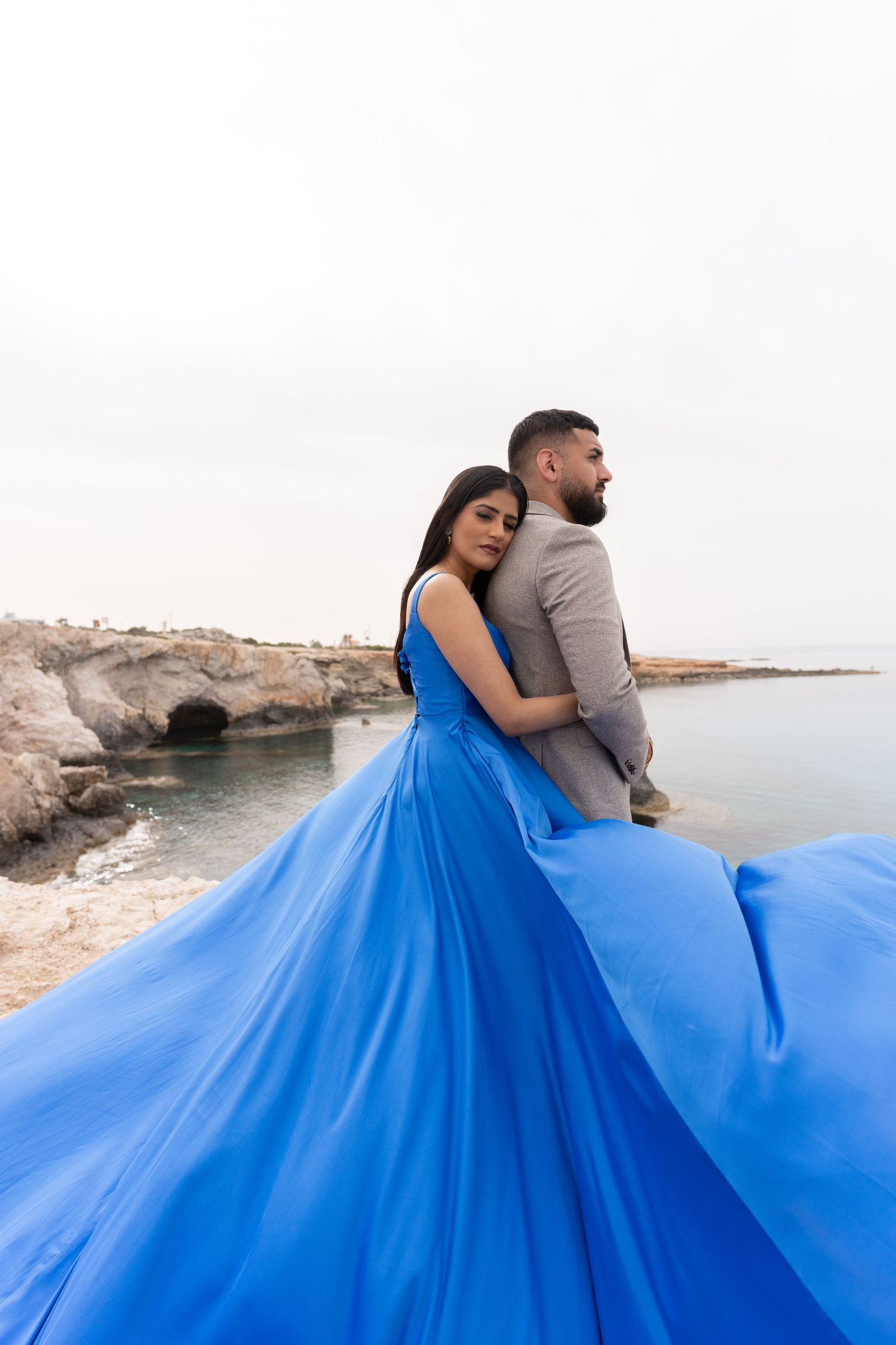 Couple embracing on rocky coast with flowing blue dress creating dramatic movement.