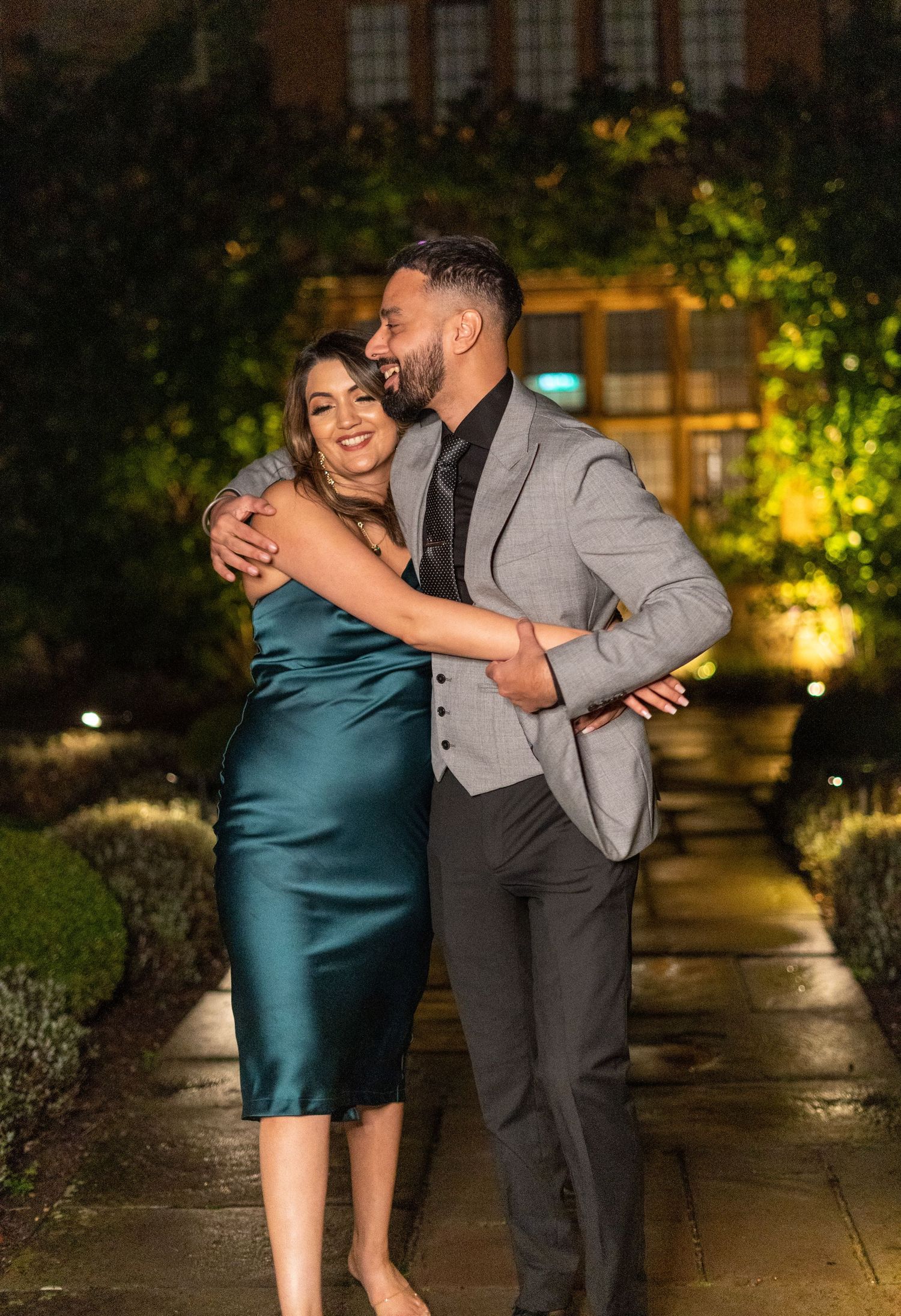 Couple embracing and laughing together at night, woman in emerald silk dress and man in grey suit.