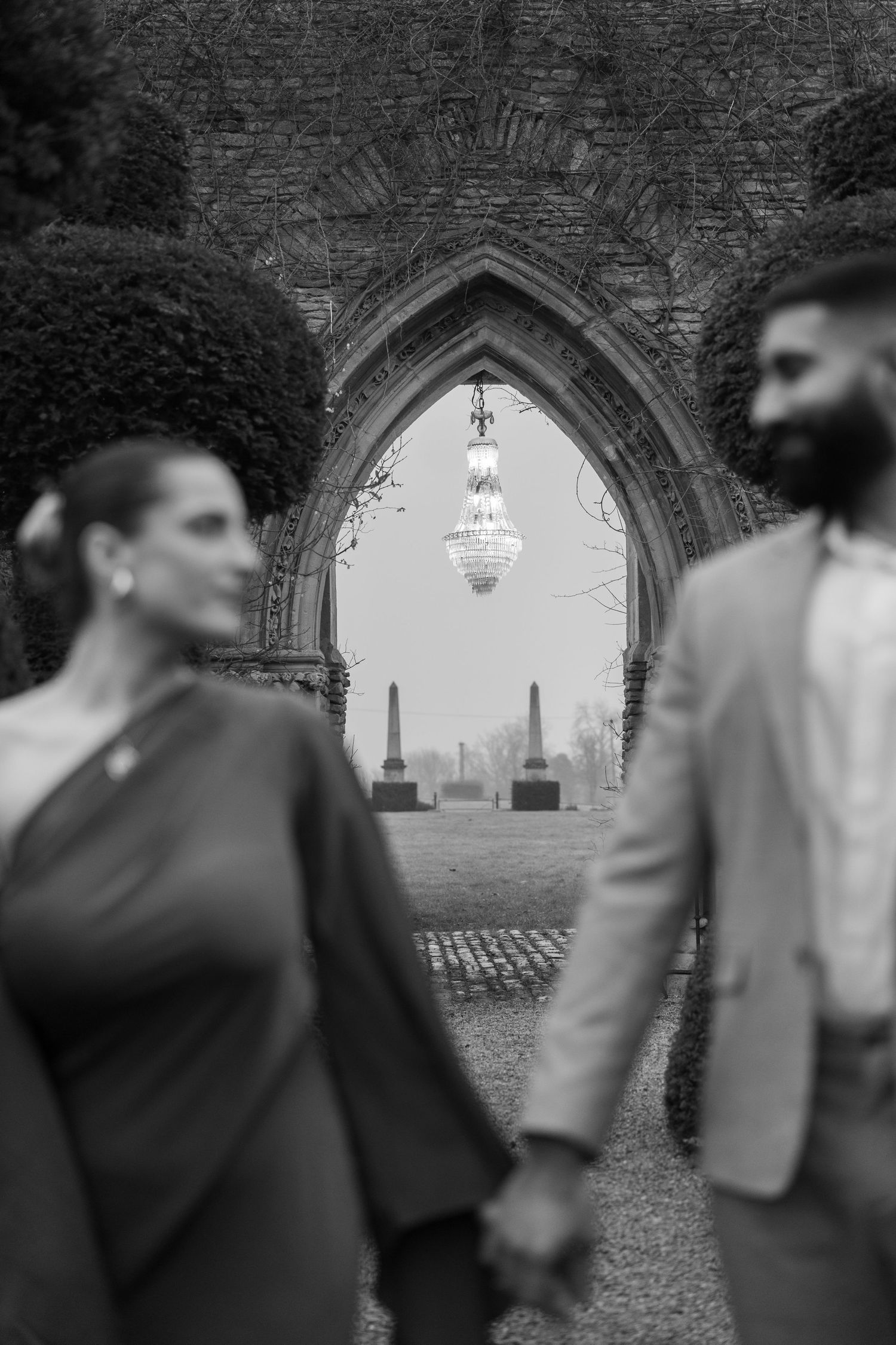 Black and white photo showing a couple holding hands through a gothic stone archway with a chandelier and cemetery in background.