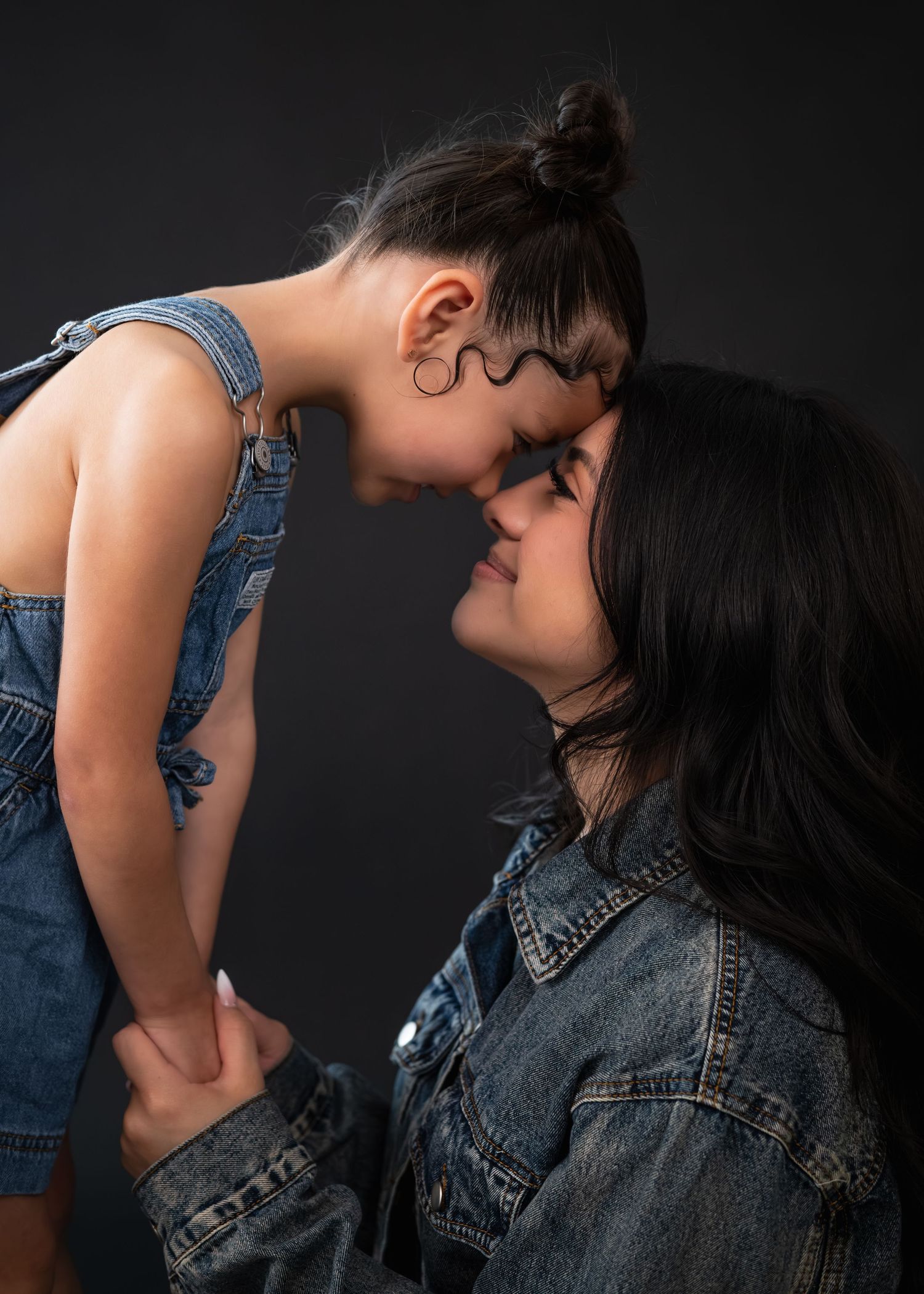 A mother and young daughter in matching denim outfits touching foreheads and smiling in a studio portrait.