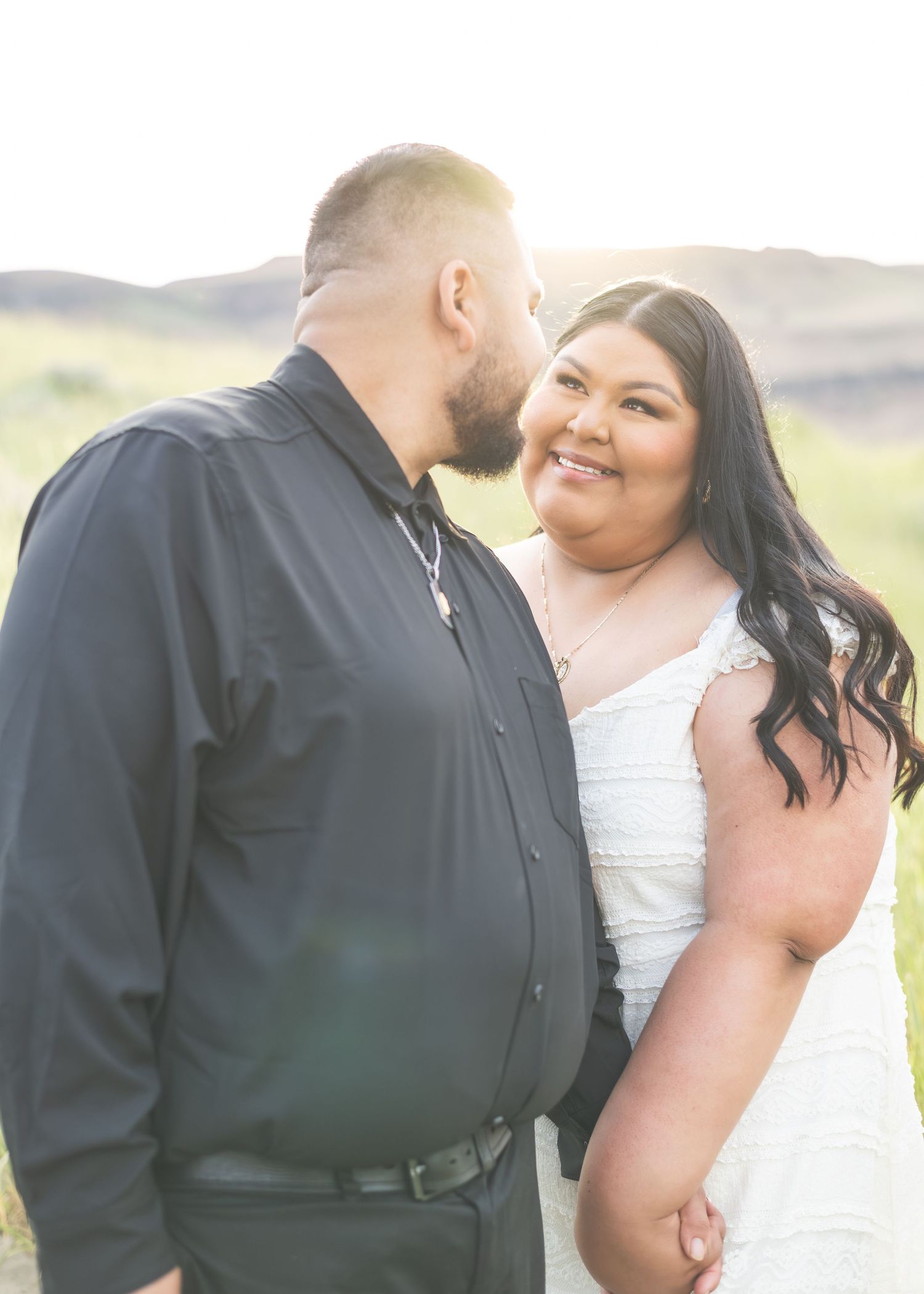 A couple shares a romantic moment outdoors at golden hour, the bride in white smiling as the groom leans in close.