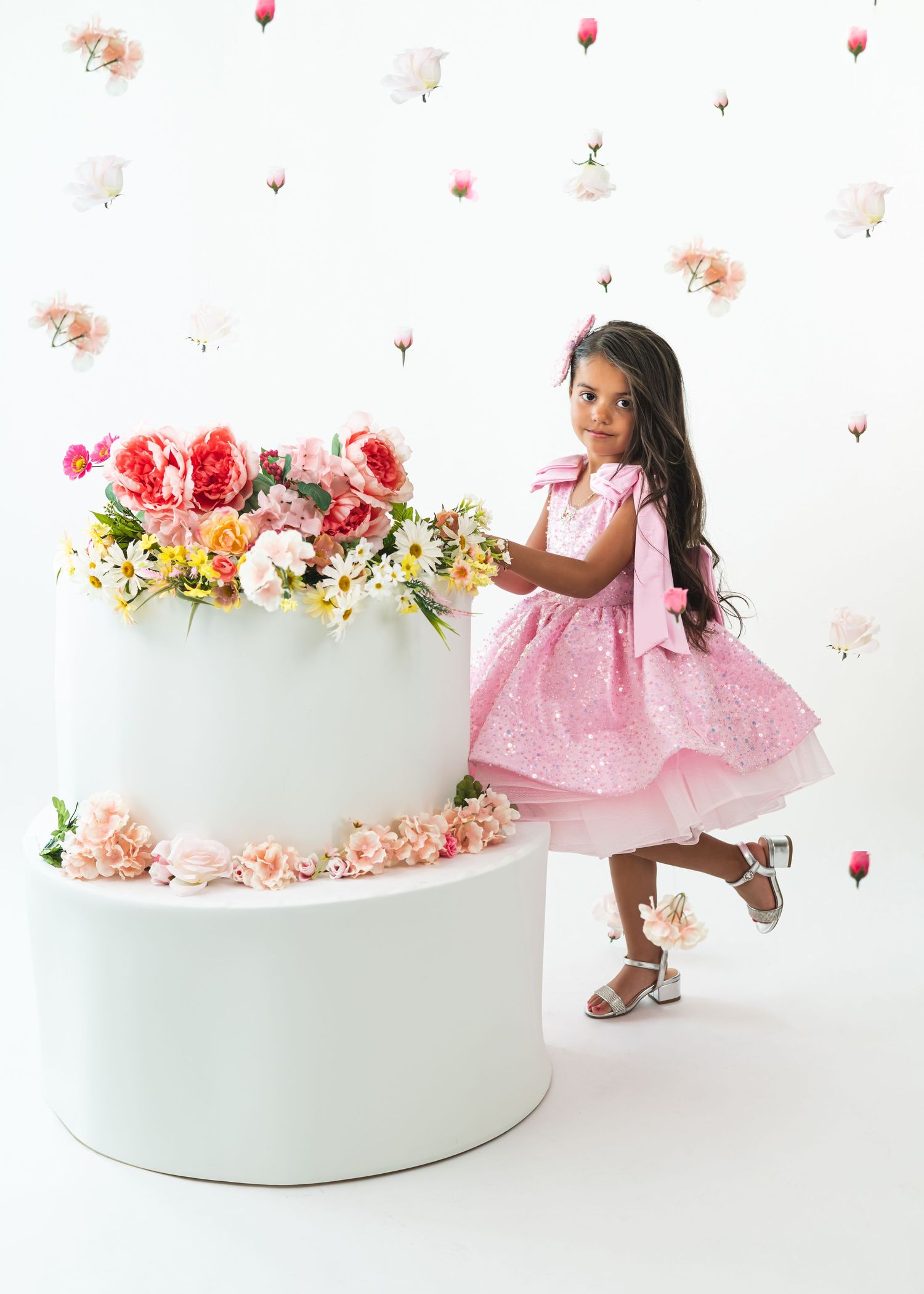 Young girl in pink dress poses beside a large white floral cake with roses on white backdrop.