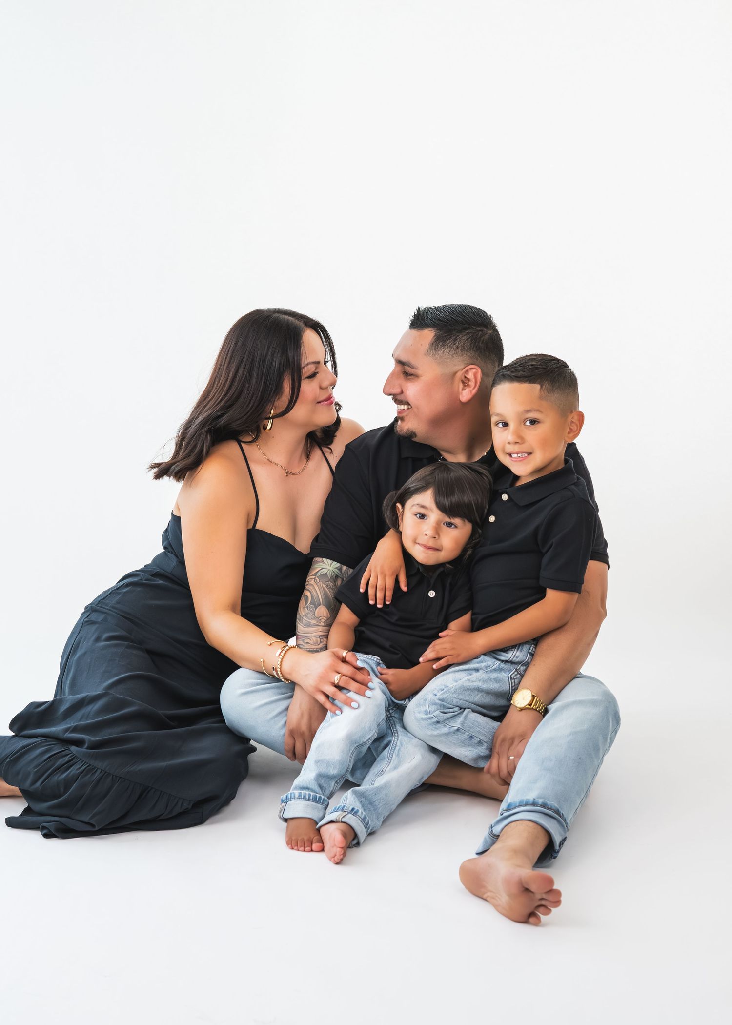 Family of four in black tops and jeans sitting together on white background, parents smiling at each other with two young boys.