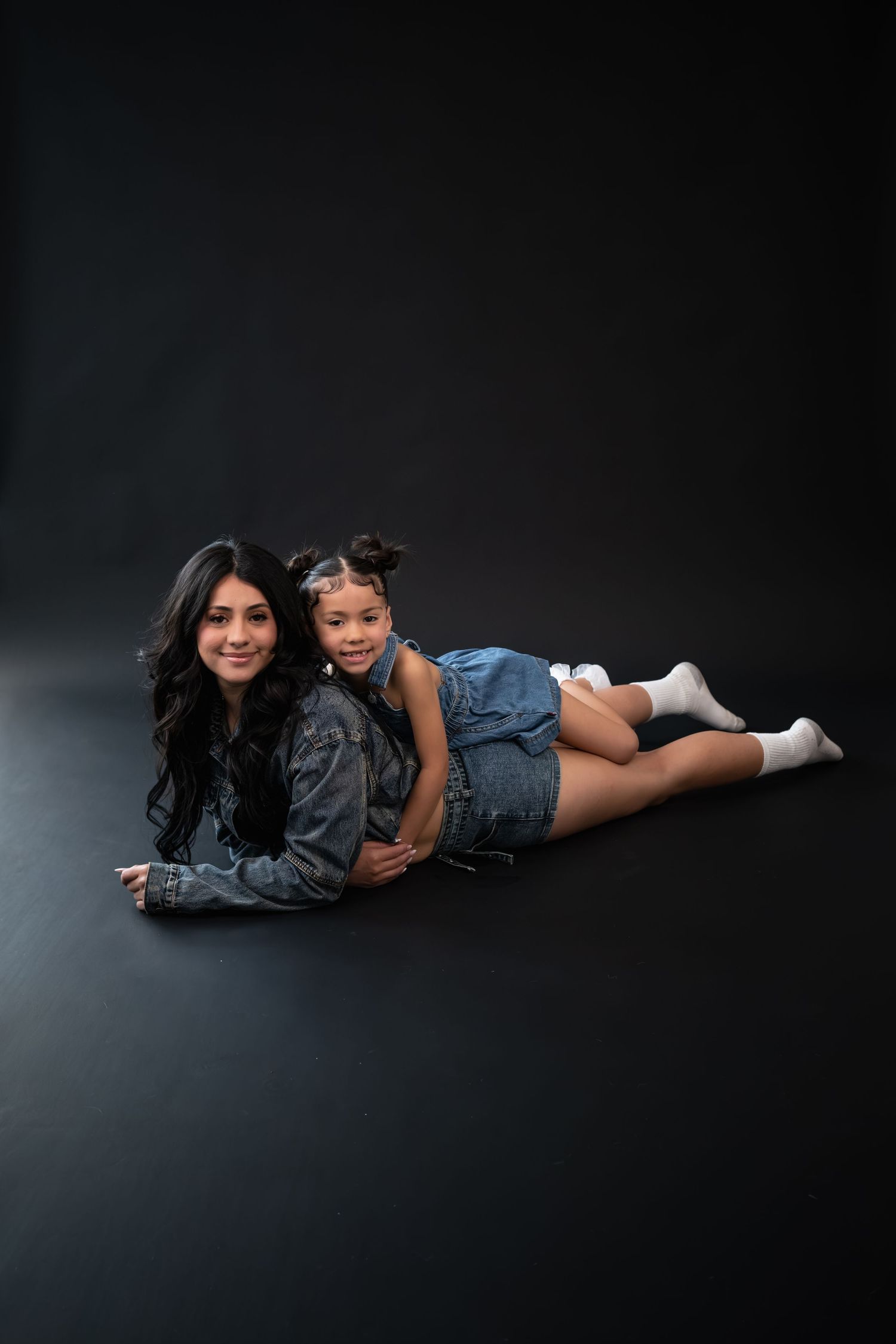 A woman and young girl in denim outfits lying on a dark floor, smiling together in a studio photo.