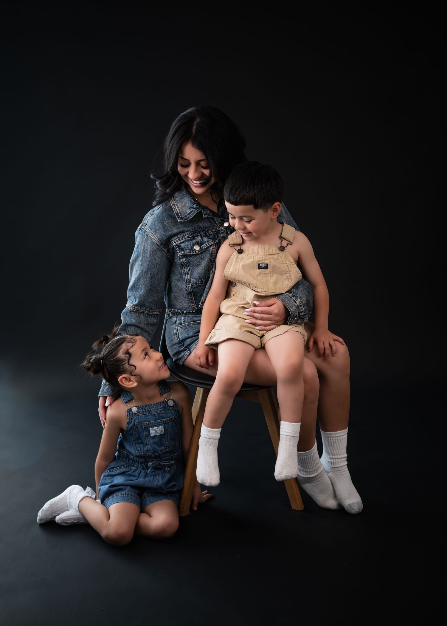 A woman in a denim jacket smiles with two young children in overalls during a studio portrait session.