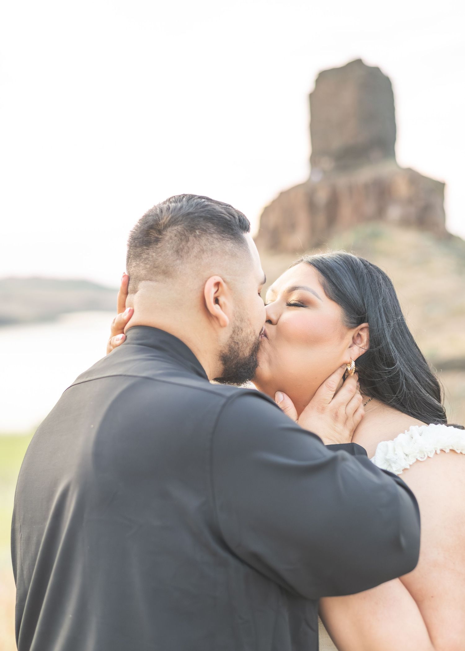 A bride and groom share a romantic kiss outdoors with a rocky formation and water in the background.