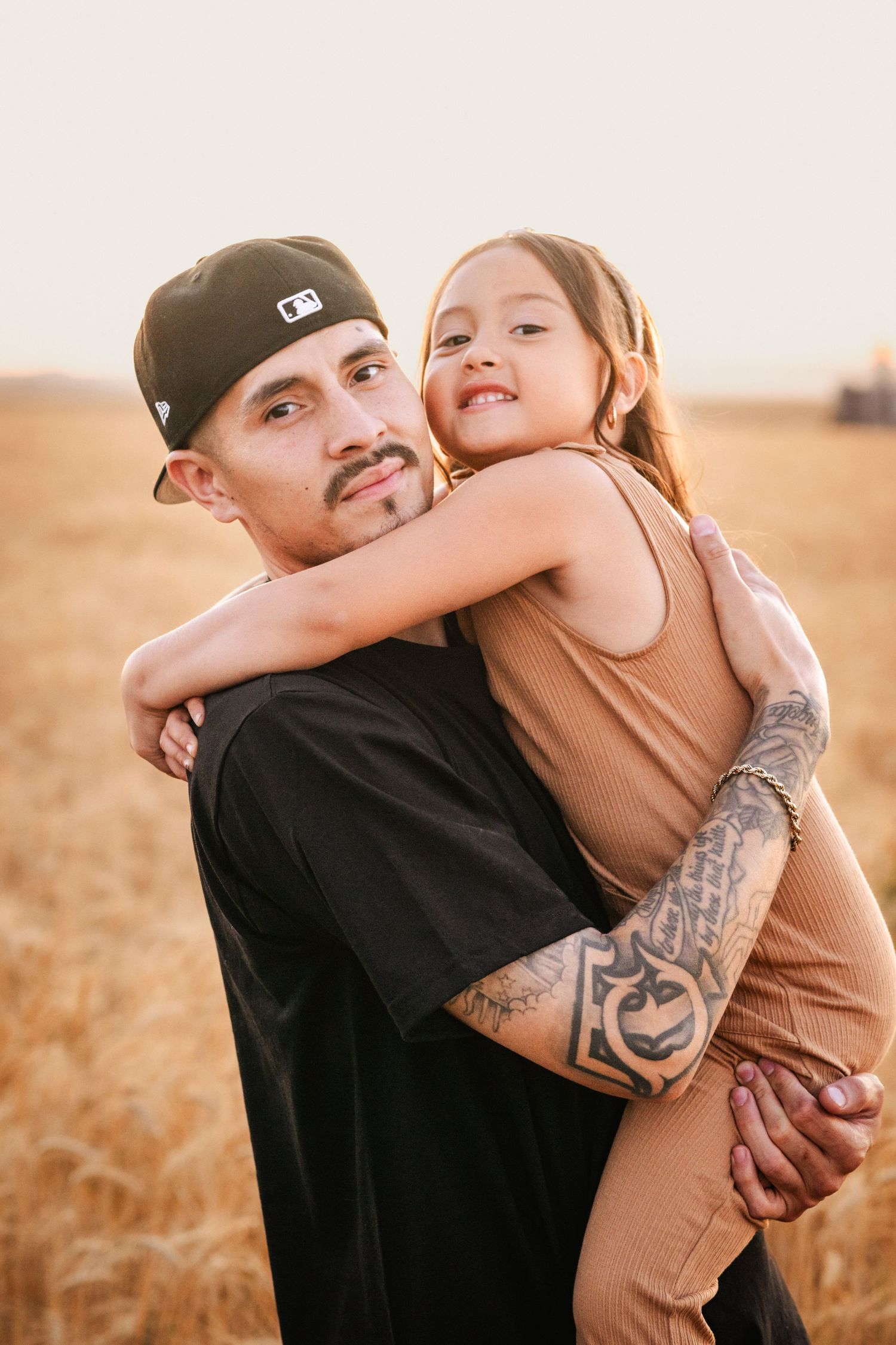 A tattooed man in a black cap holds a smiling young girl in a wheat field during golden hour.