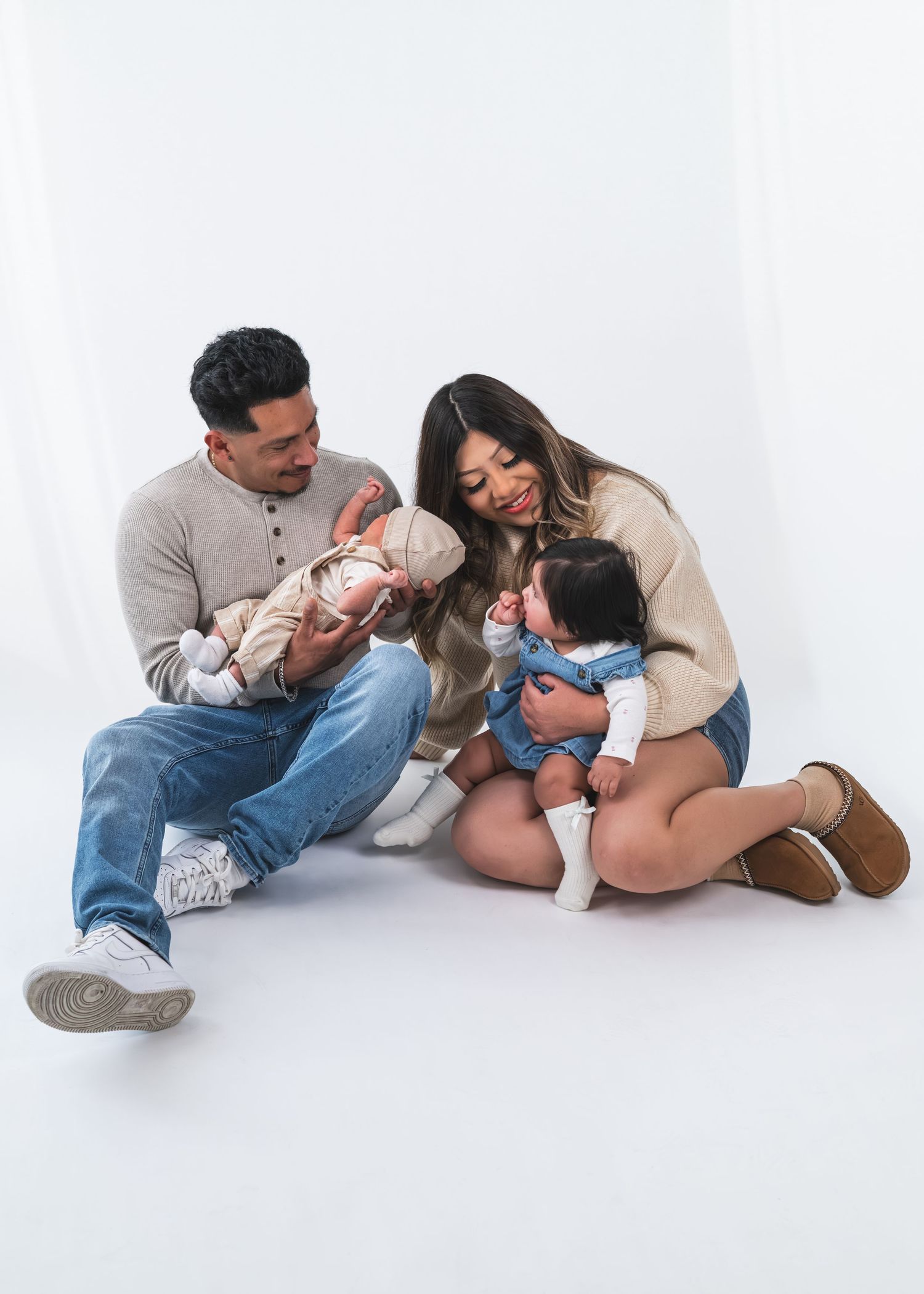 A young family sits on a white studio floor, parents lovingly gazing at their two small babies.