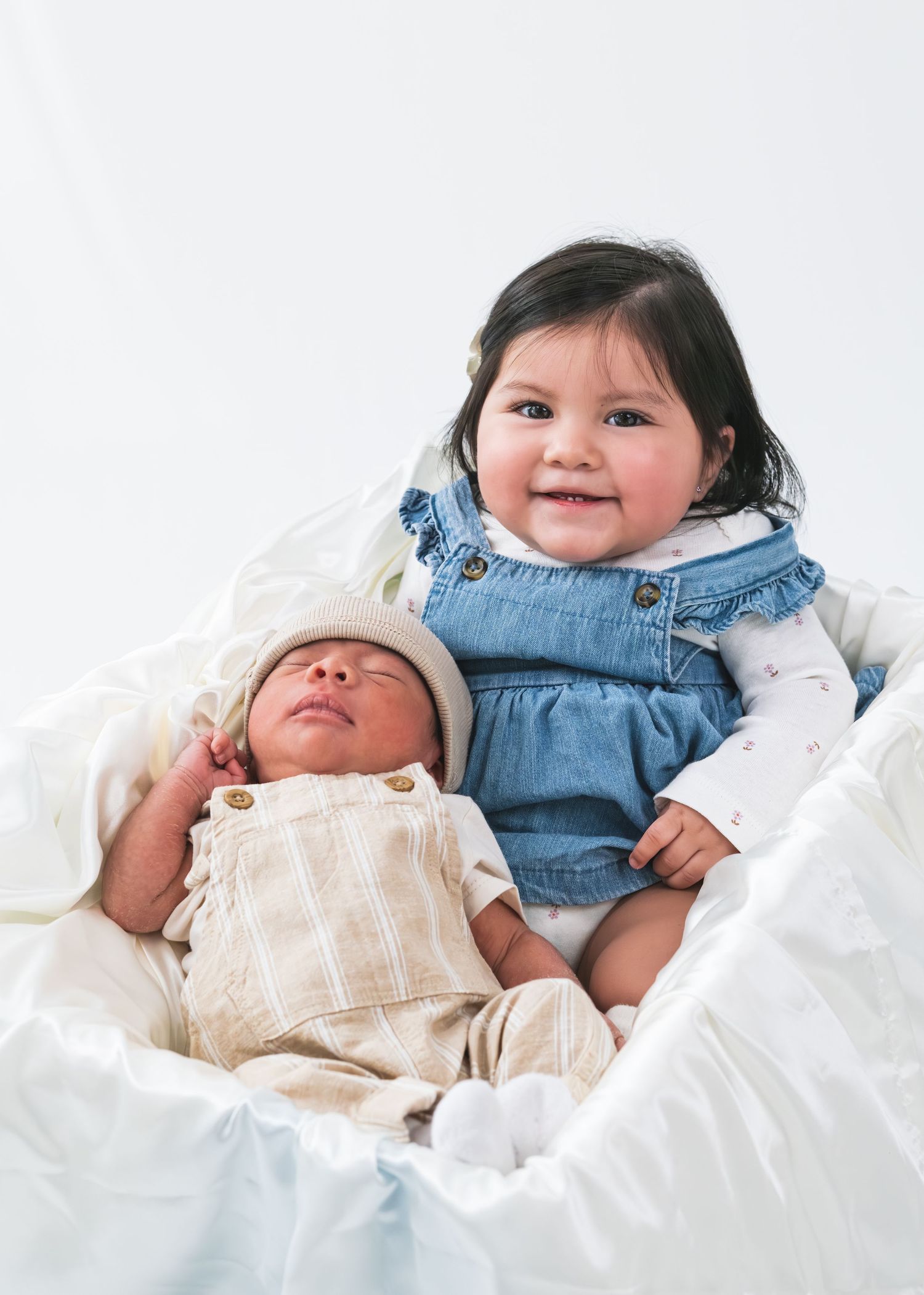 Smiling toddler girl in denim dress poses with sleeping newborn baby wrapped in white blanket.