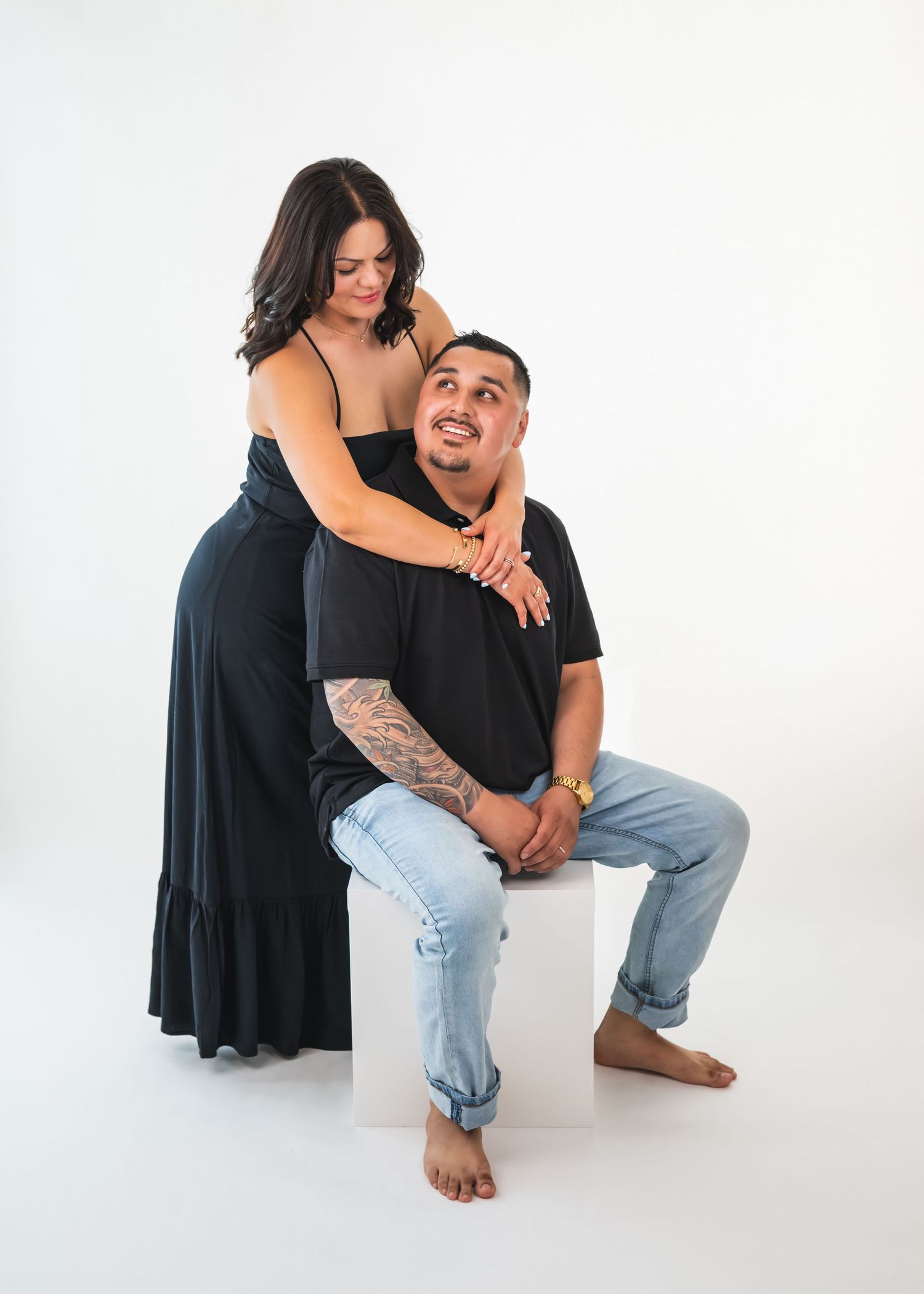 Couple in matching black outfits posing together, woman standing behind seated man, against a white studio background.